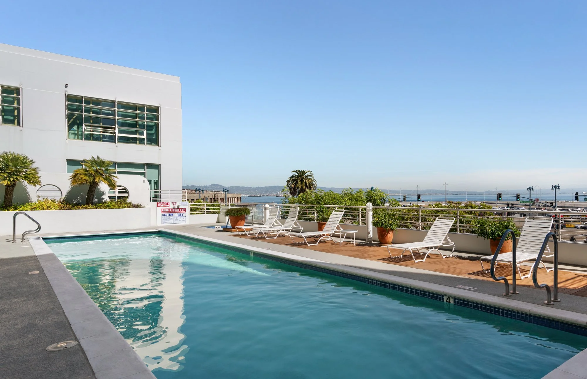 Outdoor pool area with lounge chairs and potted plants, overlooking a body of water and a partly cloudy sky.