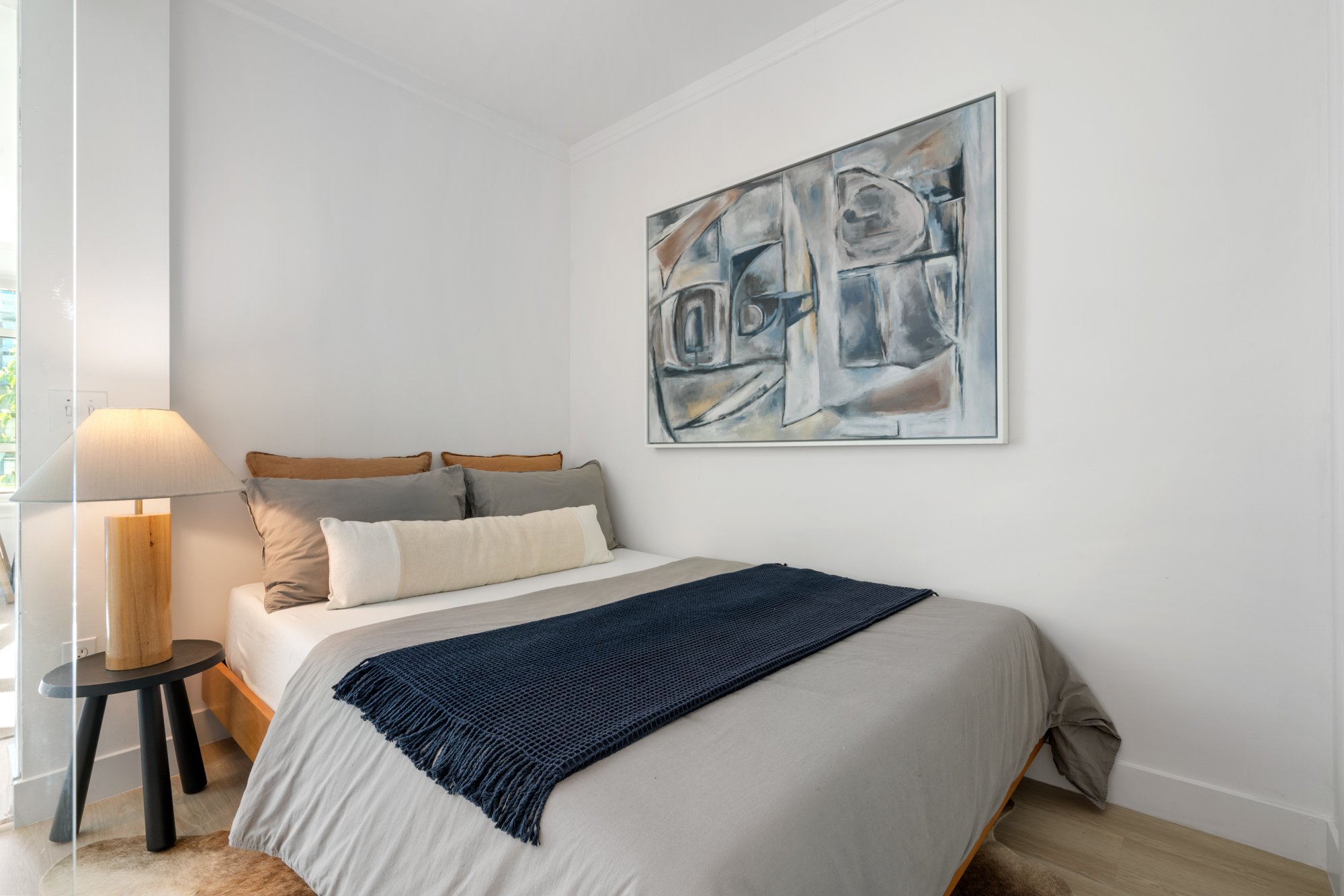 A neatly made bed with grey, beige, and brown pillows, a navy blue throw, and a beige bedspread in a modern bedroom with white walls. A black side table with a wooden lamp and a piece of abstract art above the bed.