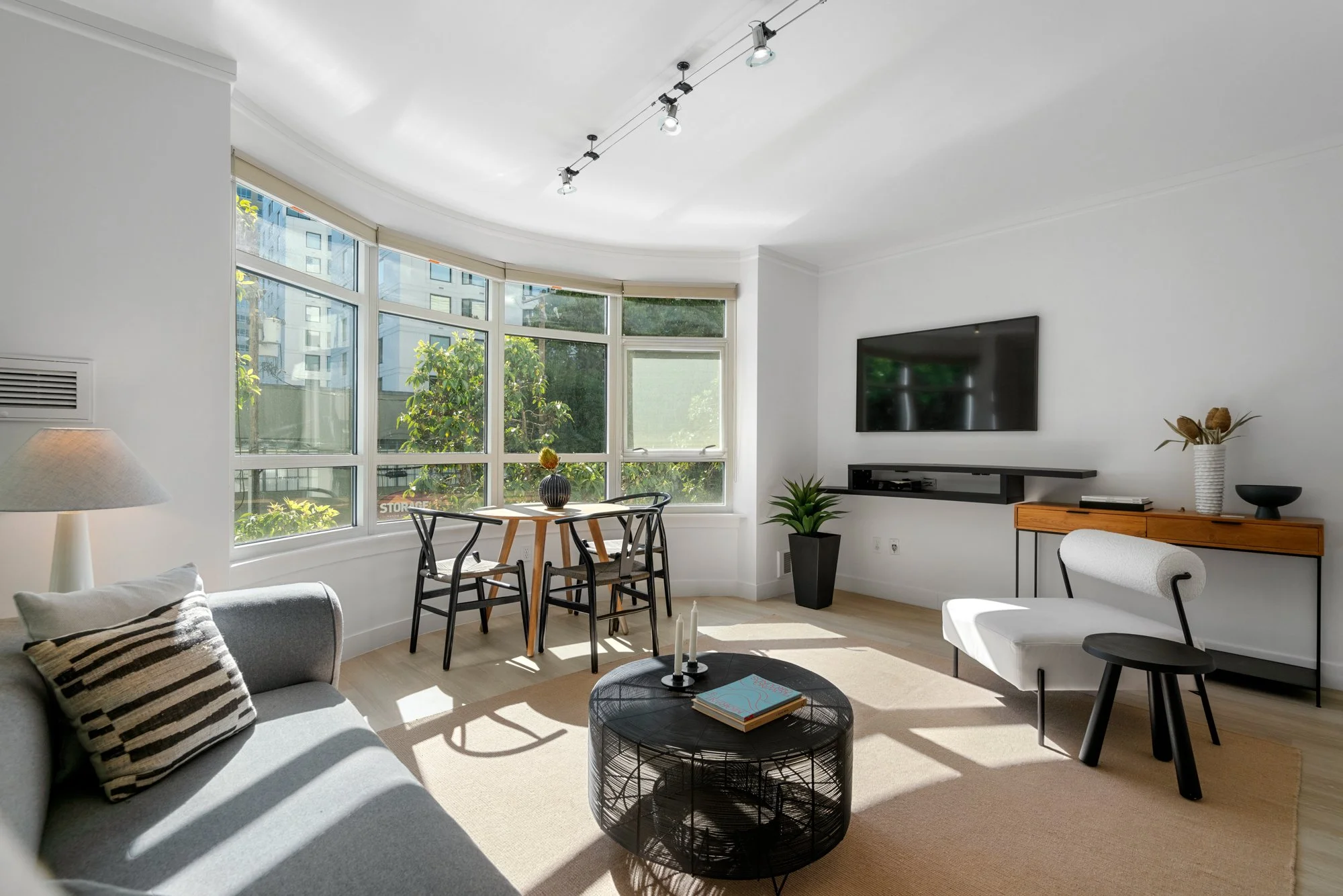 Modern living room with large curved window, gray sofa with striped pillow, black metal coffee table with books, small dining area with three chairs and a table with vase, wall-mounted TV with floating shelf, wooden sideboard with decorative objects, and potted plants, bright natural light