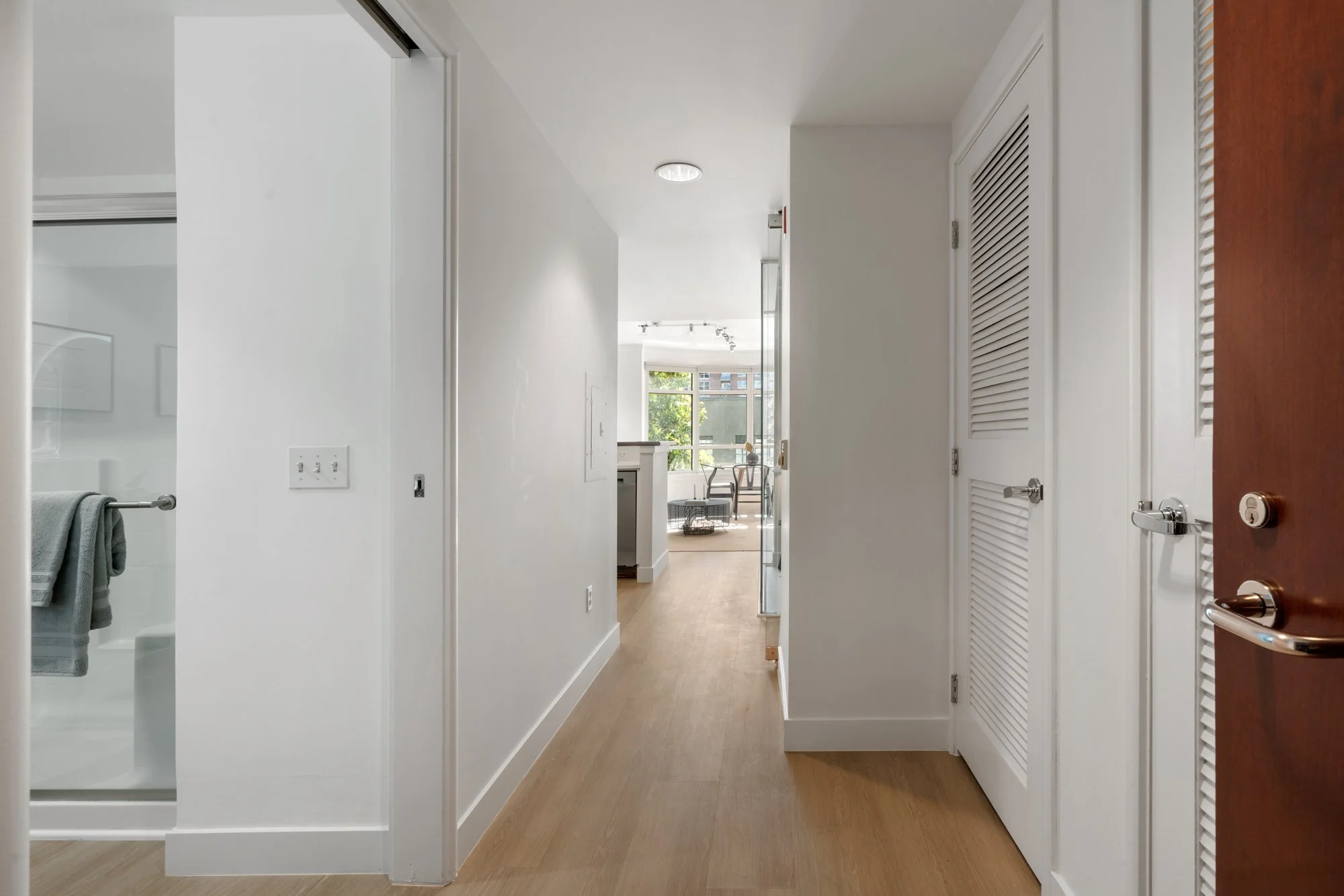 View of an apartment hallway leading to a bright living room with large windows, a small table, and chairs, with a bathroom entrance on the left side of the hallway.