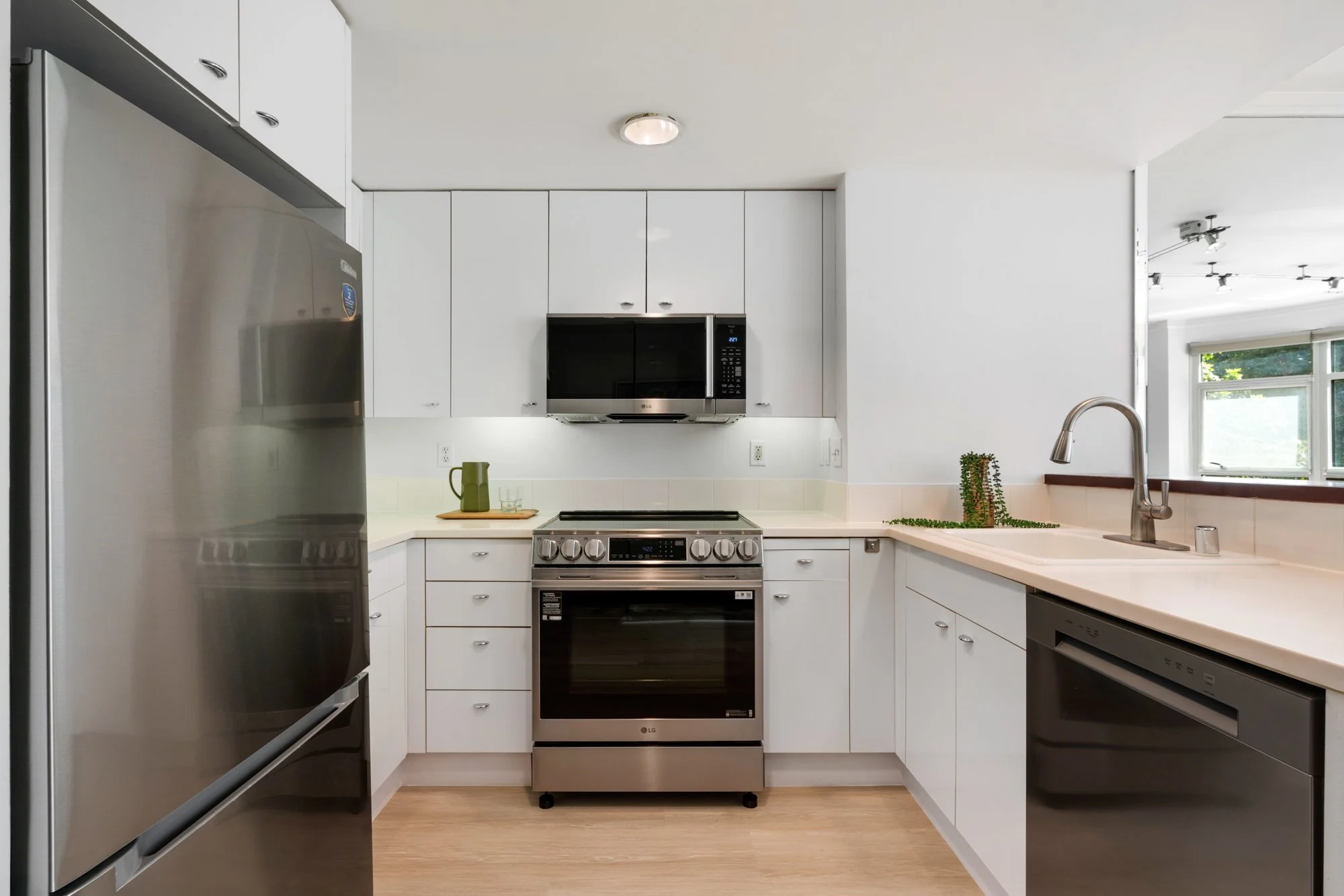 Modern kitchen with white cabinets, stainless steel appliances including refrigerator, oven, microwave, and dishwasher, and a double sink near a window.