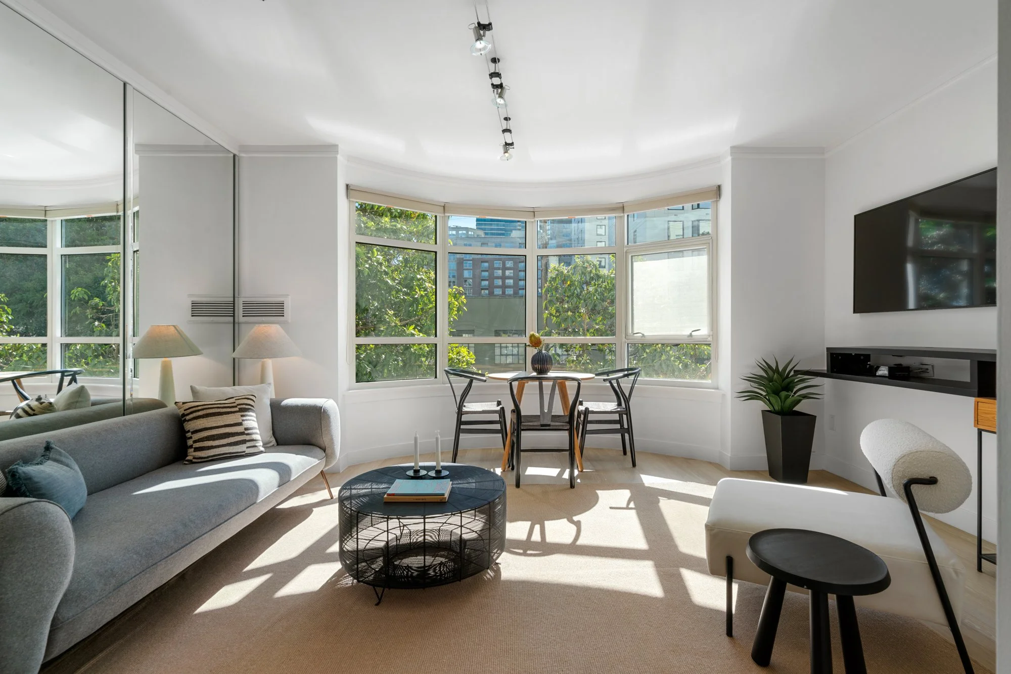 Living room with large bay windows, a gray sofa with throw pillows, a round black coffee table, white walls, modern furniture, a wall-mounted TV, and indoor plants.