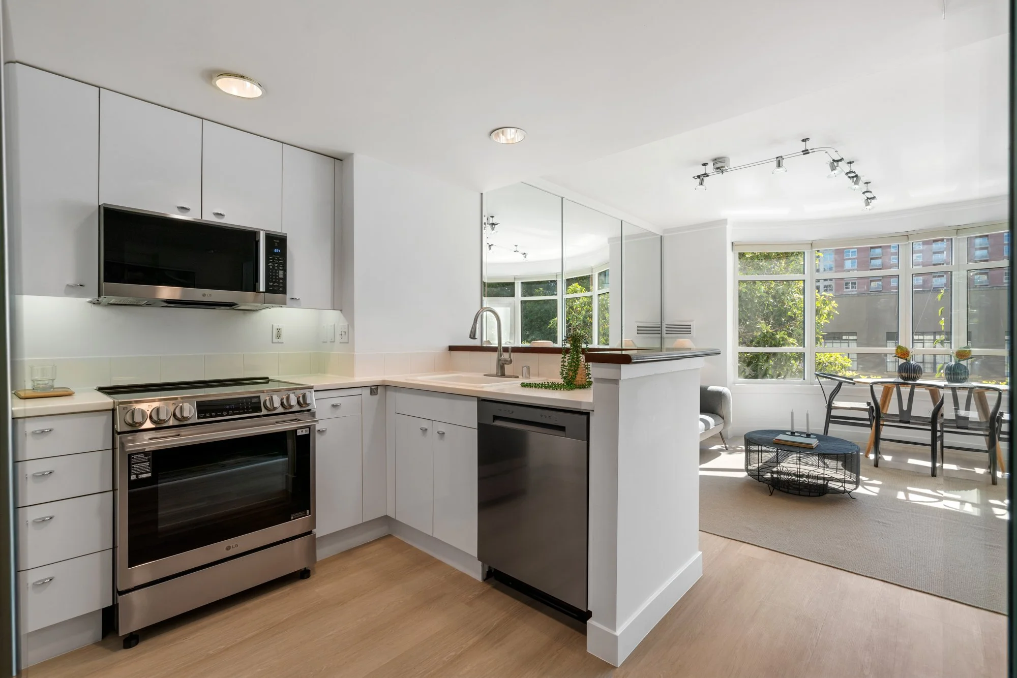 Modern kitchen with white cabinets, stainless steel stove, microwave, and dishwasher; open living space with large windows, dining table, and sitting area, decorated with plants.