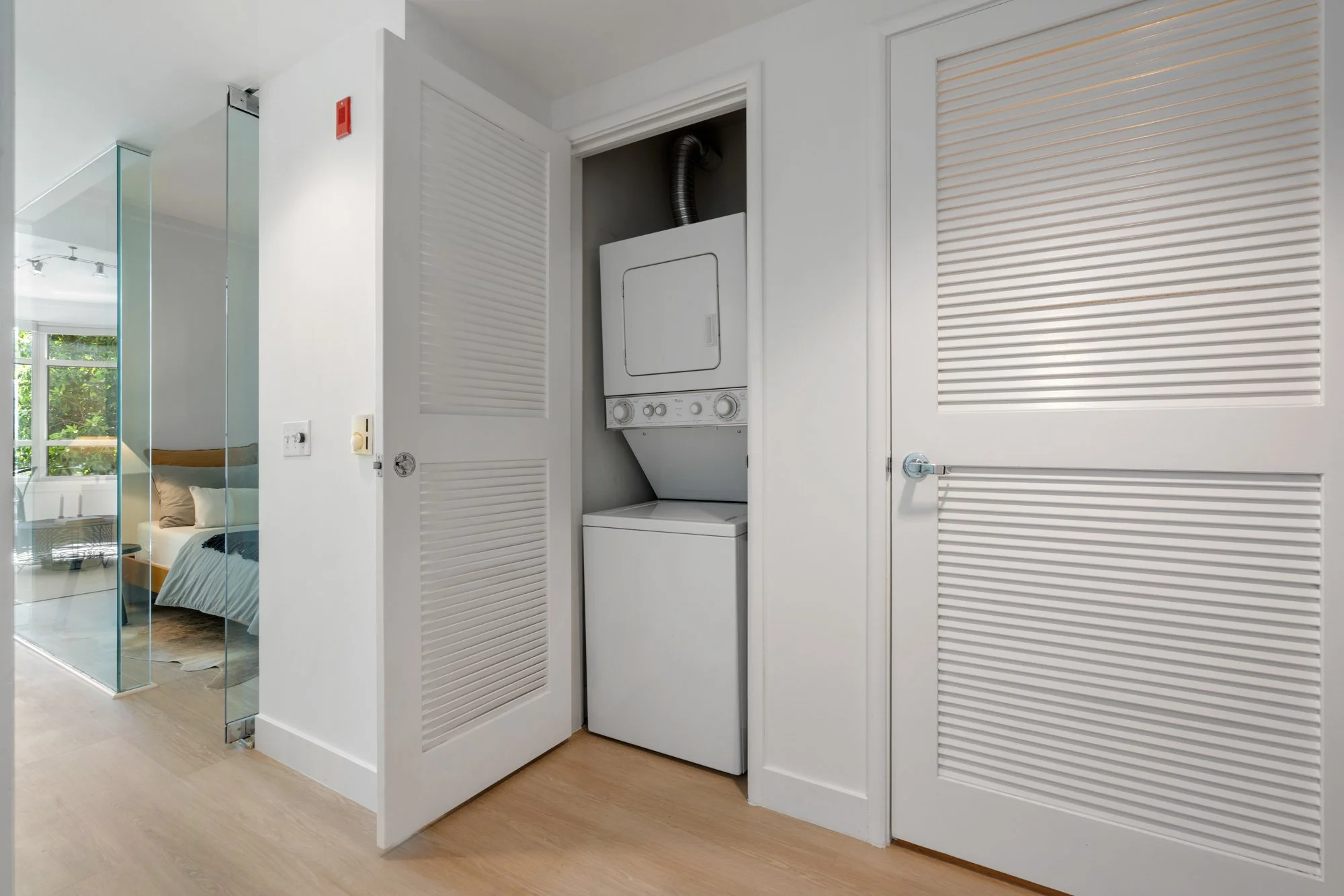 A laundry closet with a stacked washer and dryer behind white louvered bifold doors in a modern apartment.
