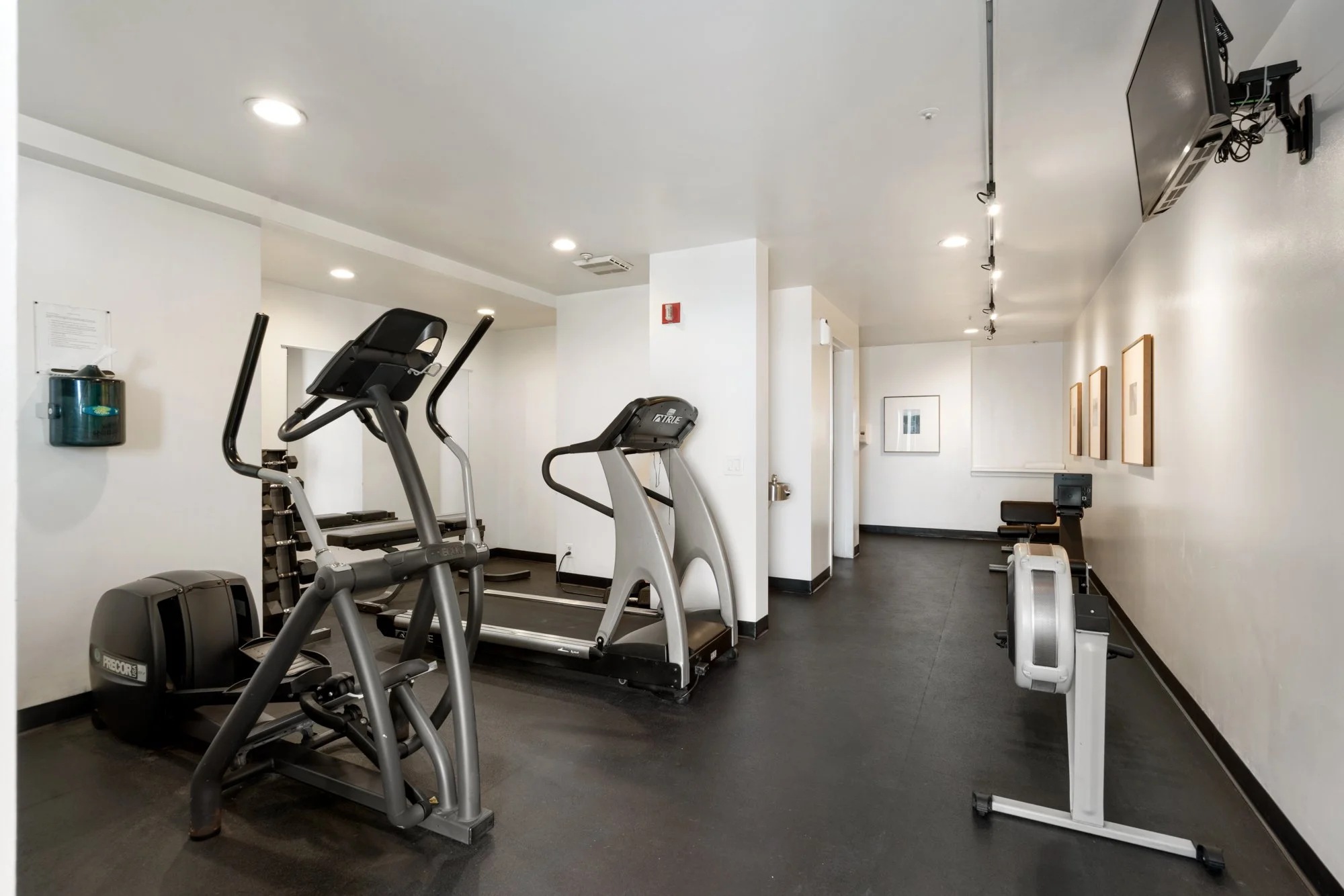 Empty gym room with exercise equipment including a treadmill, elliptical machine, and rowing machine, with white walls and black flooring.