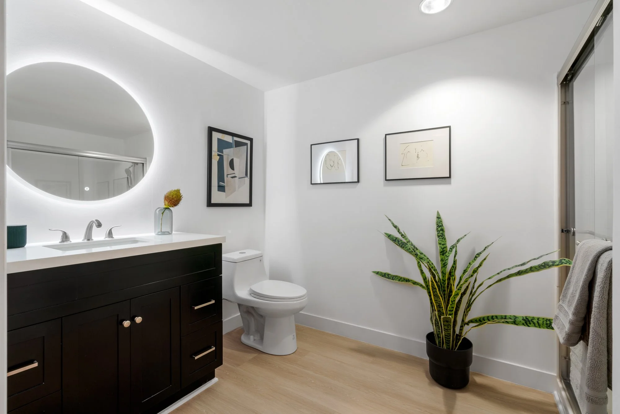Modern bathroom with a round backlit mirror, black vanity with white countertop, toilet, framed artworks, potted snake plant, and a towel on a hook.
