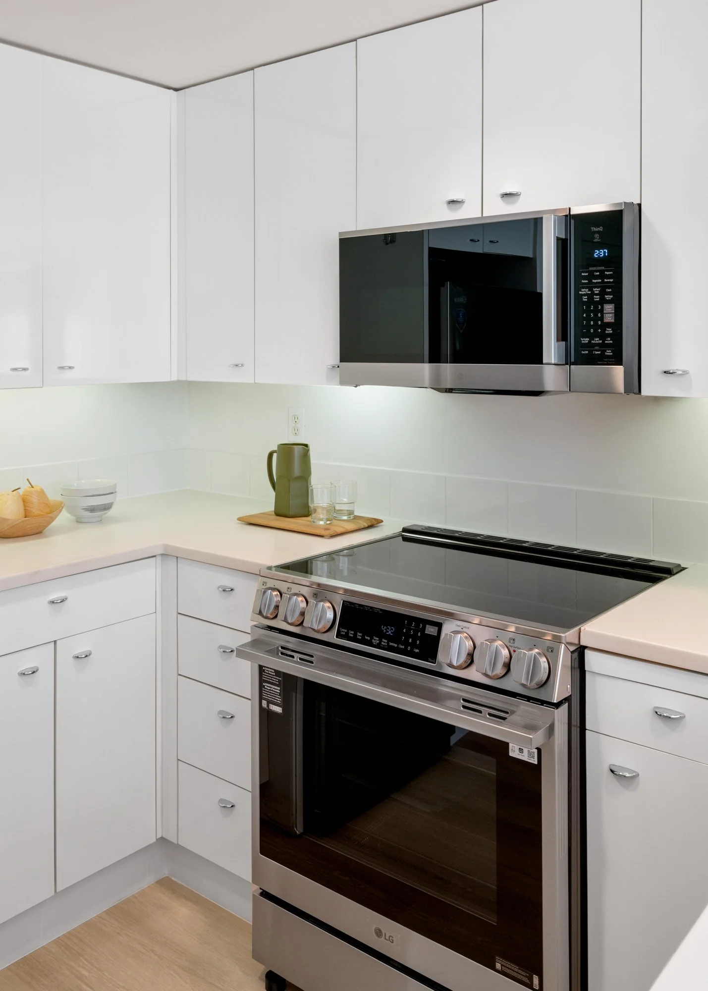 Modern kitchen with white cabinets, stainless steel microwave and oven, beige countertop, and light wood floor.
