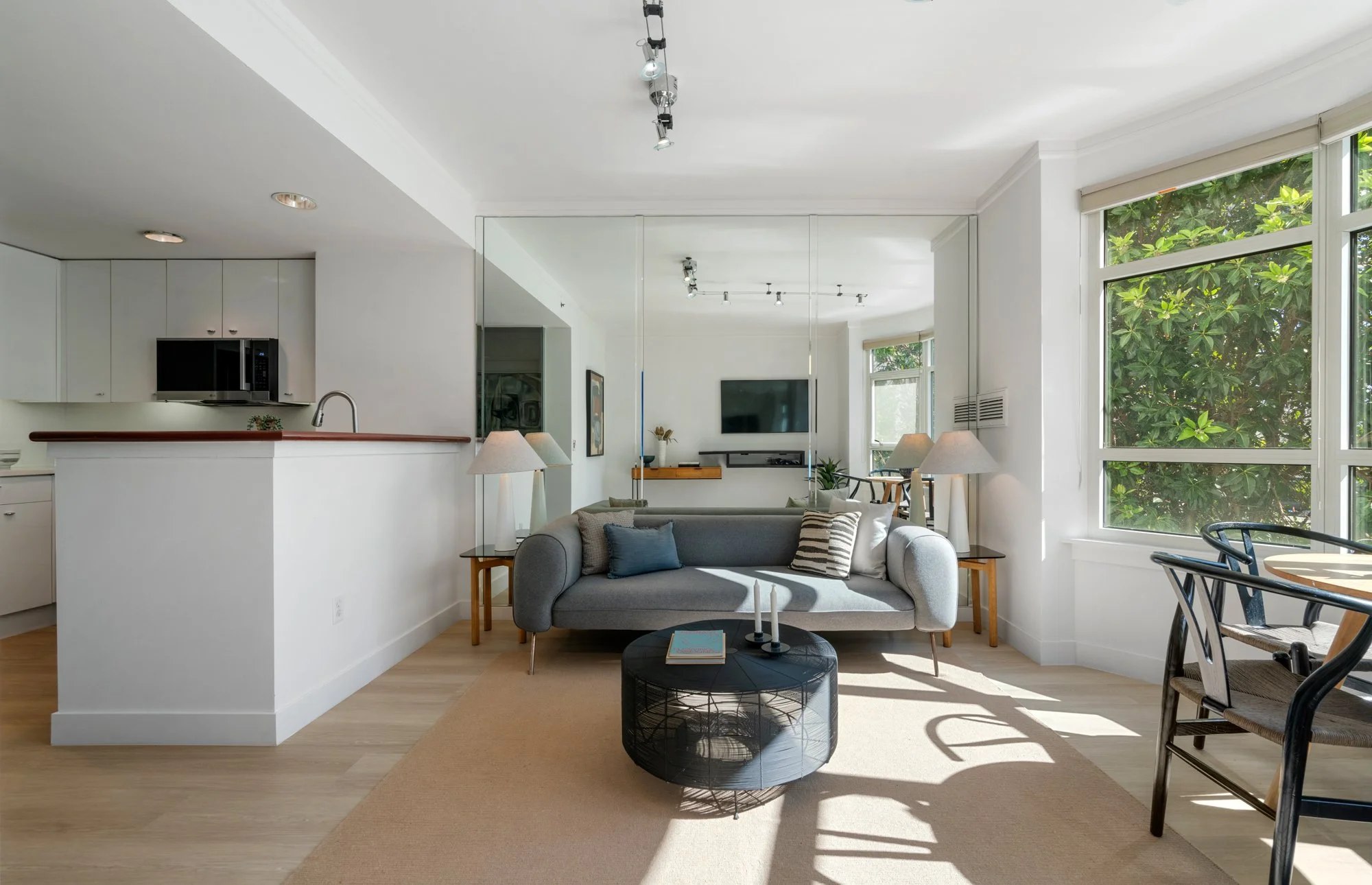 Living room with gray sofa, black coffee table, side tables with lamps, large windows with greenery outside, and a white kitchen in the background.