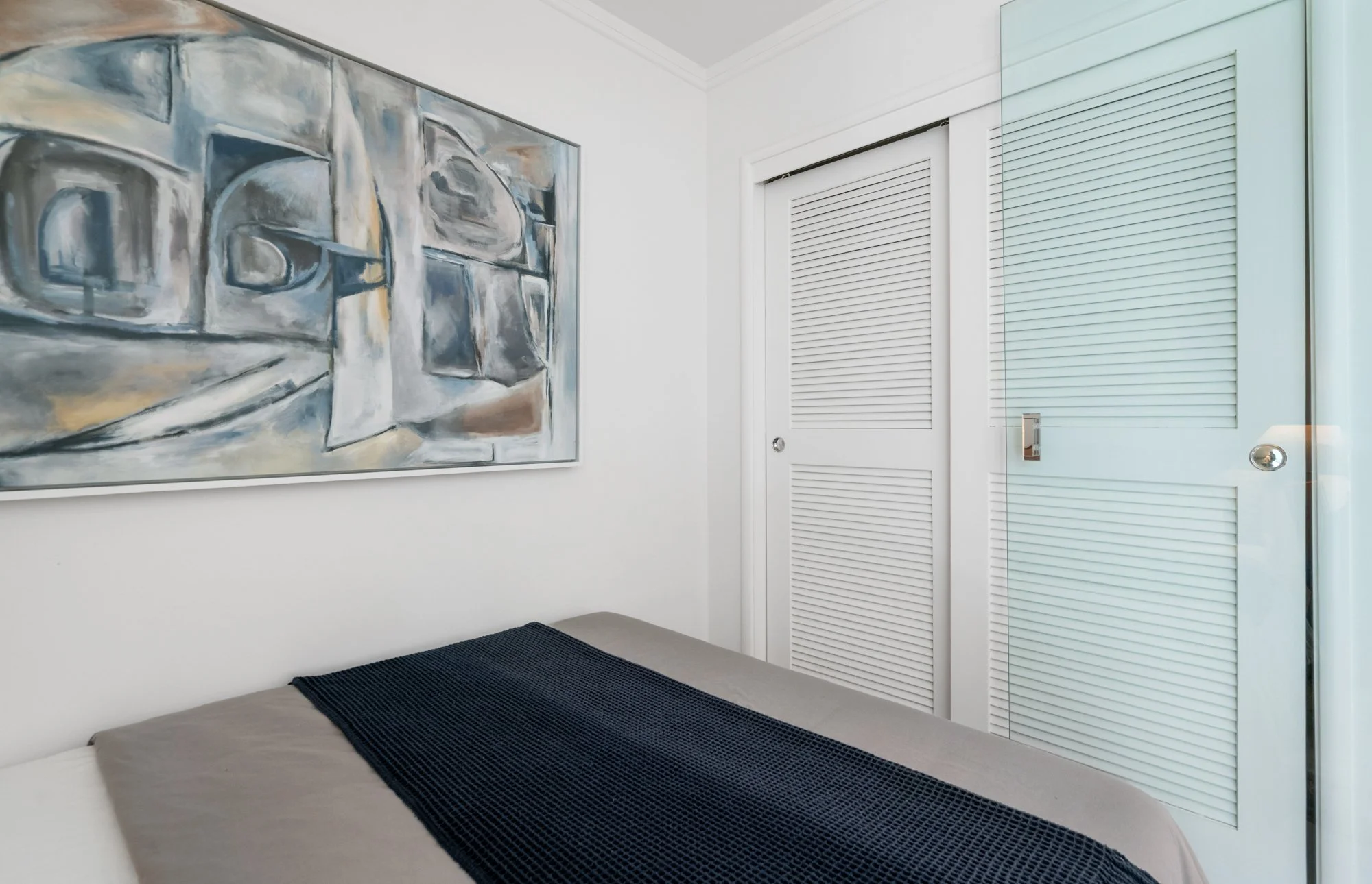 A bedroom with a grey bed, a blue blanket, abstract artwork on the wall, a white closet door, and a window with blue blinds.