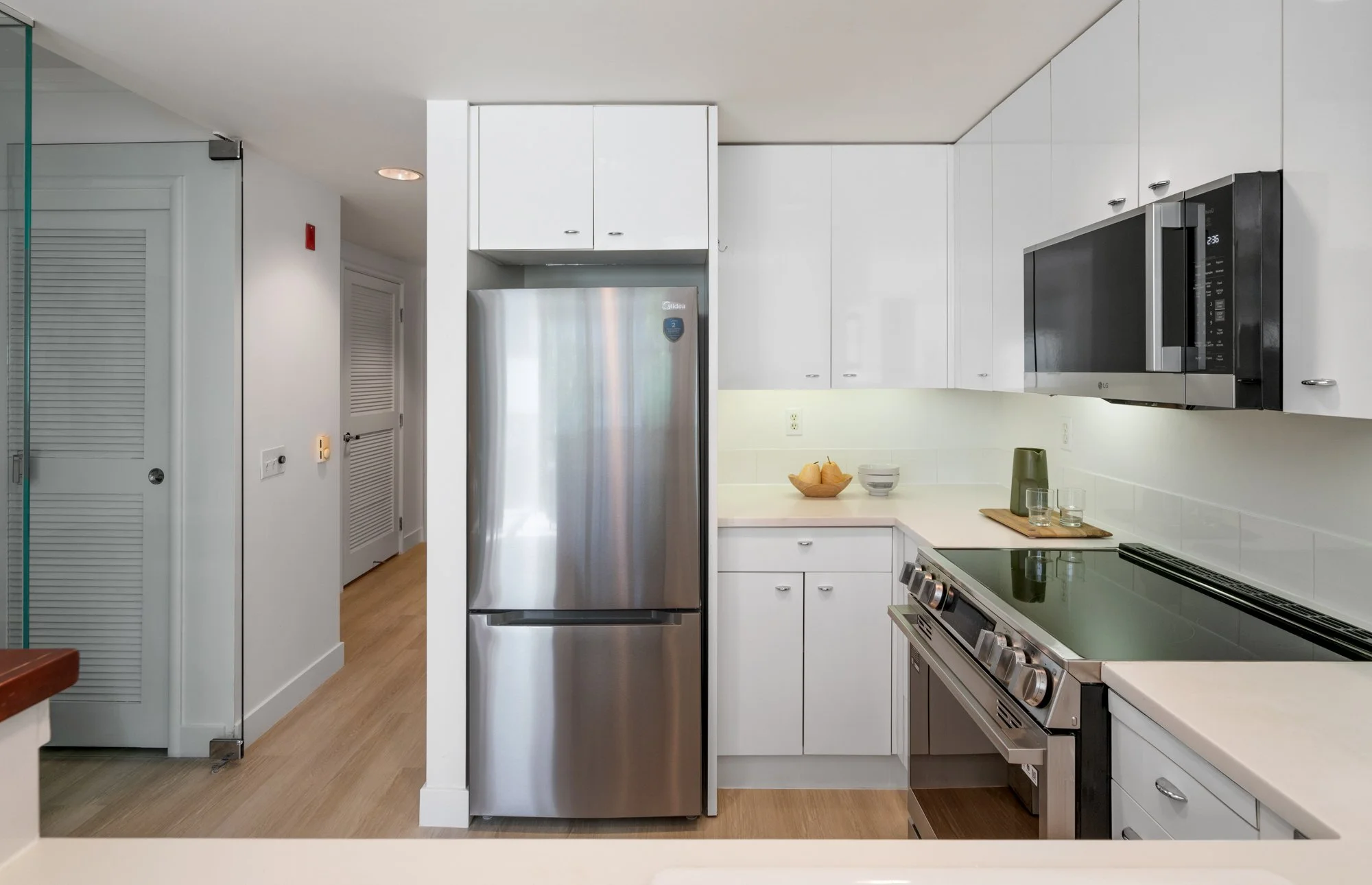 Modern kitchen with stainless steel refrigerator, microwave, and stove, white cabinets, light wood flooring, and a small countertop with bowls and glasses.