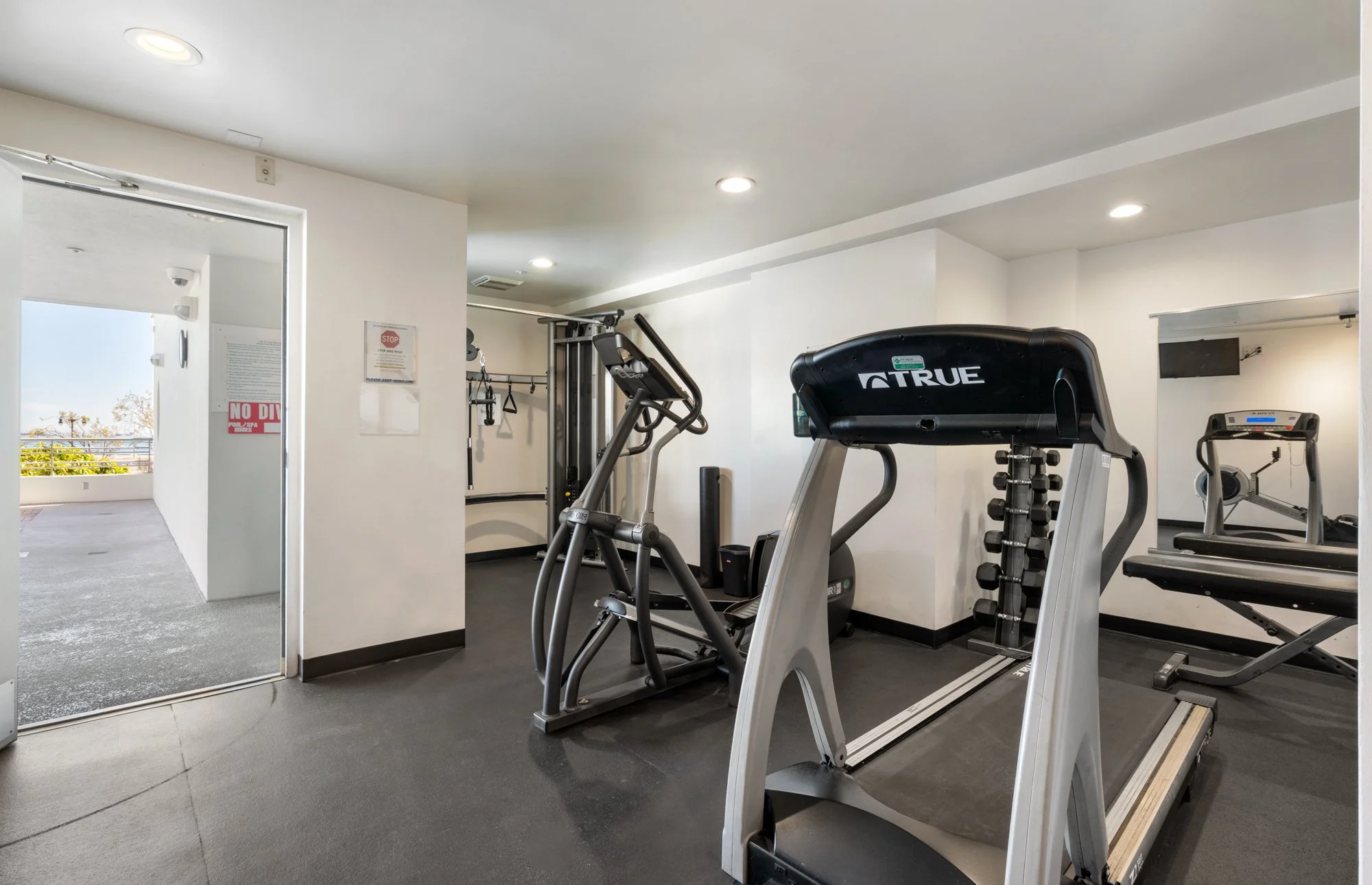 Indoor gym with treadmill, elliptical machine, dumbbell rack, and exercise equipment, near a doorway leading to a balcony.
