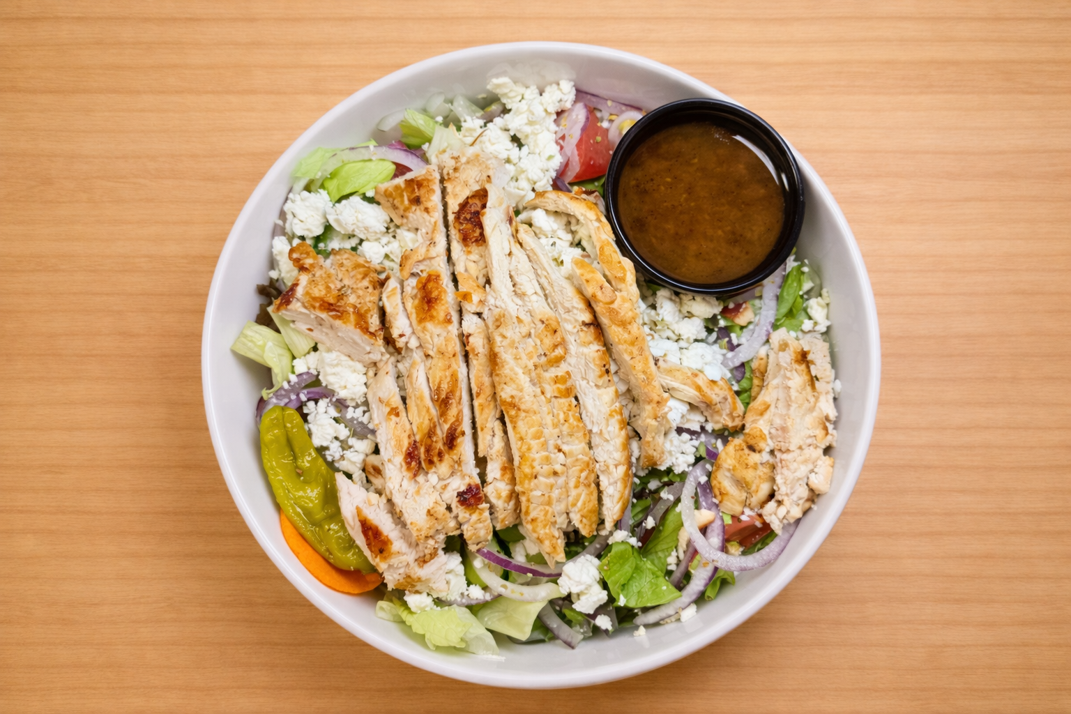 Salad with grilled chicken slices, mixed greens, shredded cheese, sliced red onion, tomato, green pepper, and a side of dressing in a small black bowl.