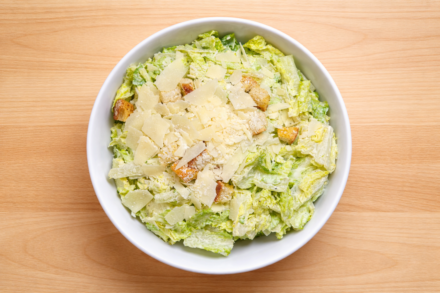 Caesar salad with romaine lettuce, grated Parmesan cheese, croutons, and Caesar dressing in a white bowl on a wooden surface.