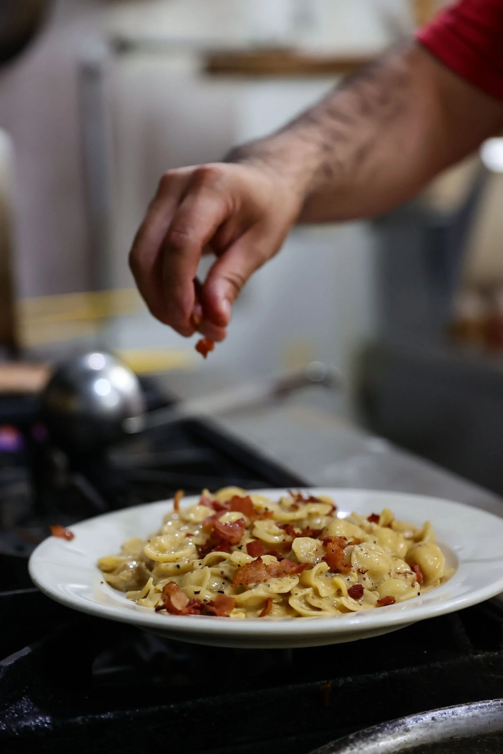 A chef sprinkling seasoning on a plate of cooked pasta with bacon bits.