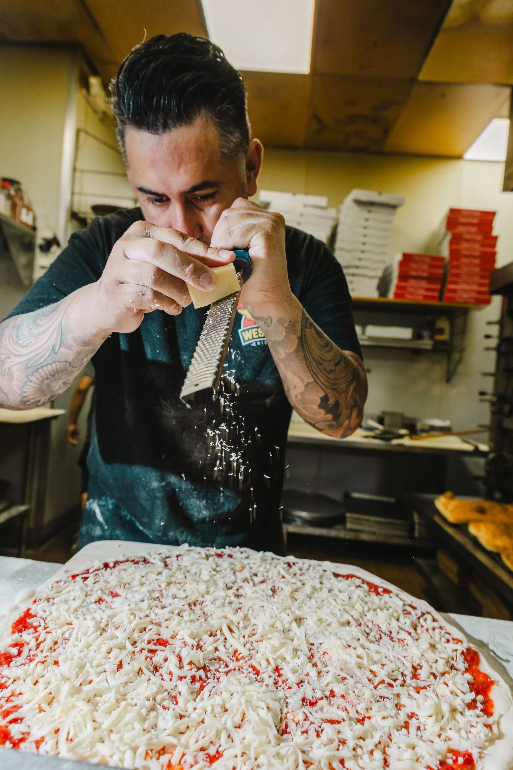 A man with tattoos grating cheese over a large pizza in a pizzeria kitchen.