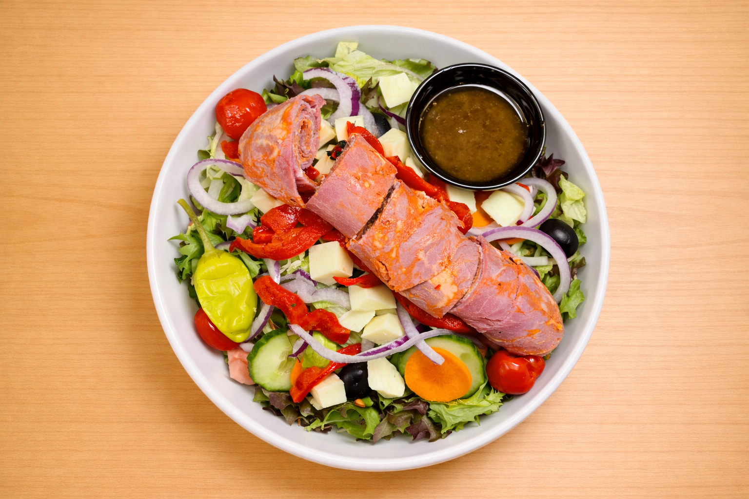 Salad with mixed greens, cherry tomatoes, cucumber, red onion, black olives, cheese cubes, red chili peppers, and a slice of ham or luncheon meat, served with a small bowl of dressing on a white plate on a wooden surface.