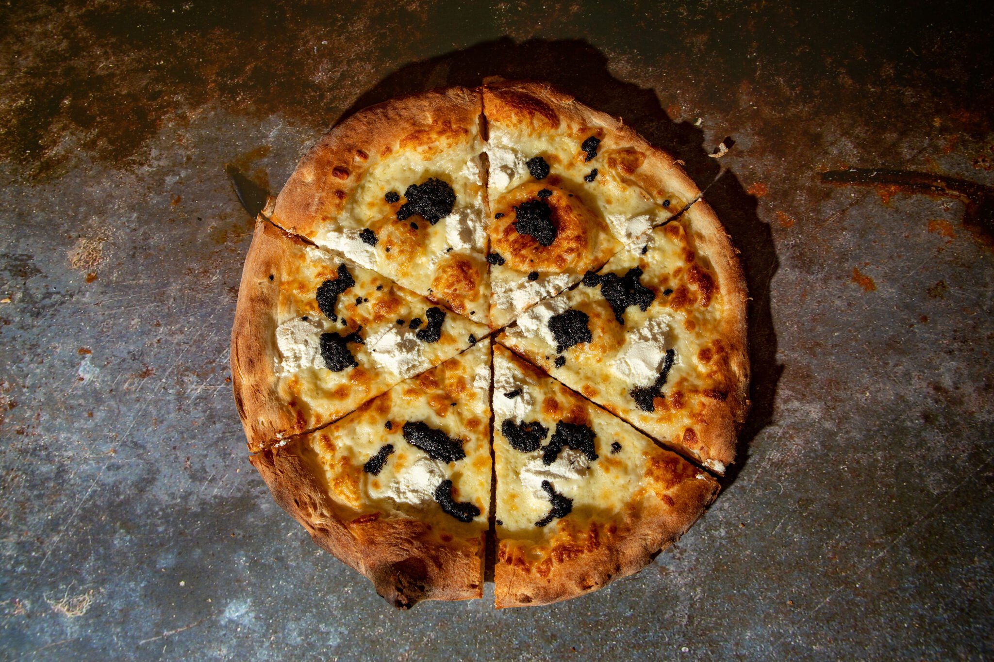 A partially sliced pizza with cheese and black toppings on a dark, textured surface.