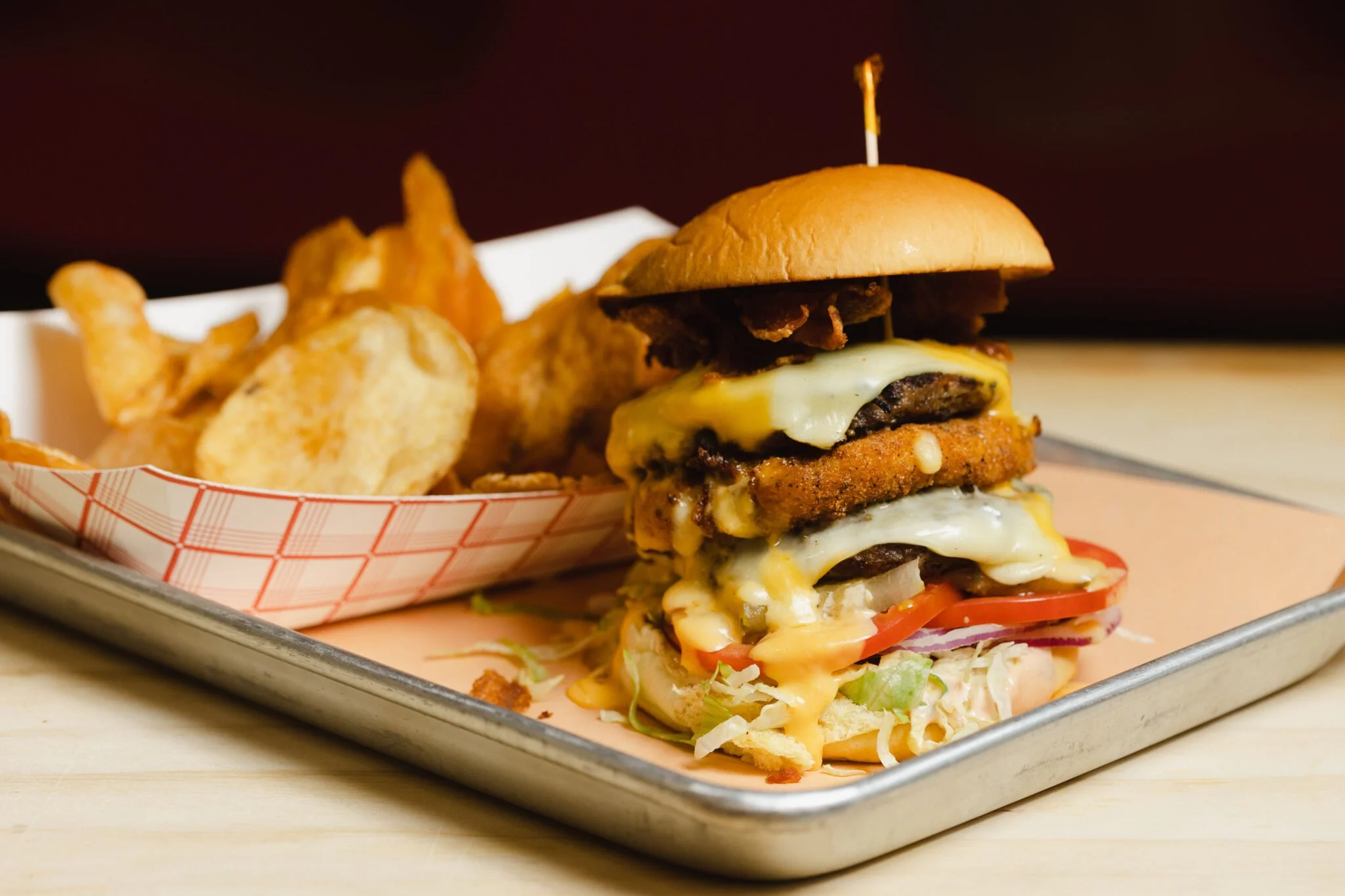 A cheeseburger with multiple beef patties, melted cheese, tomato slices, lettuce, and onions, served with potato chips on a tray.
