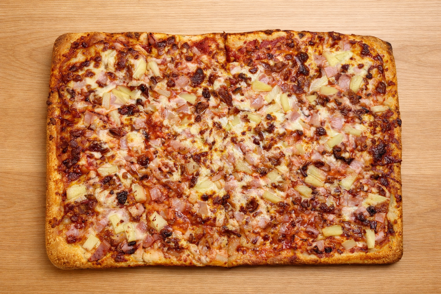 Rectangular pizza with cheese, ham, pineapple, and drizzled sauce on a wooden surface.