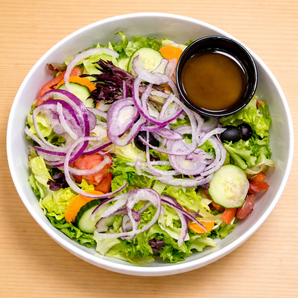 A white bowl filled with a colorful salad containing lettuce, cucumber, tomato, purple onion, black olives, and a small container of salad dressing on a light wooden surface.