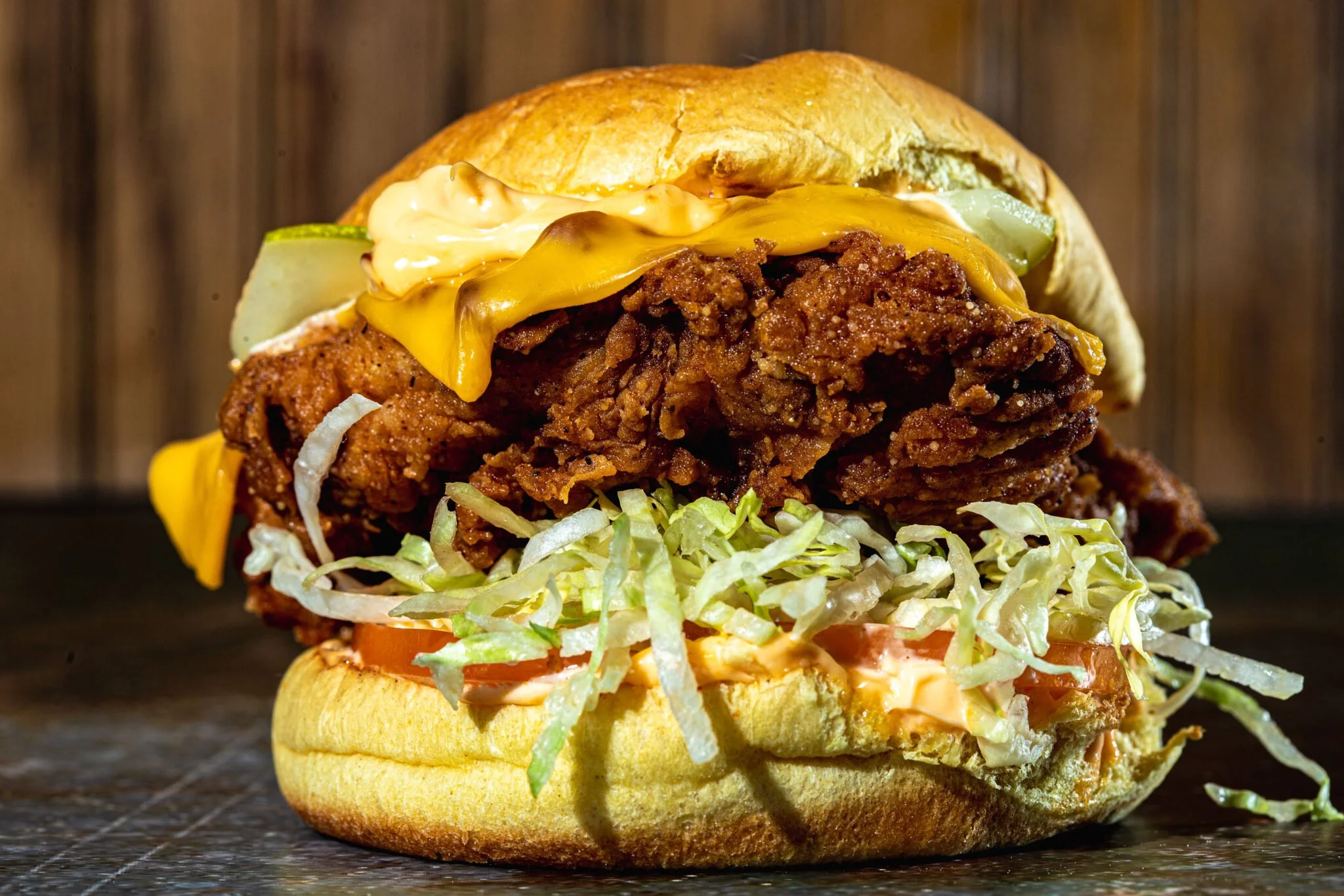 Close-up of a sandwich with fried chicken, cheddar cheese, pickles, lettuce, tomato, and sauce in a bun on a dark surface.