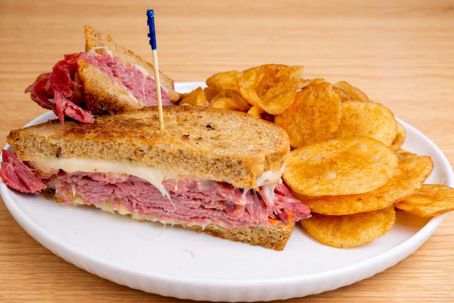 Sandwich with corned beef, melted Swiss cheese, and rye bread, served with potato chips on a white plate.