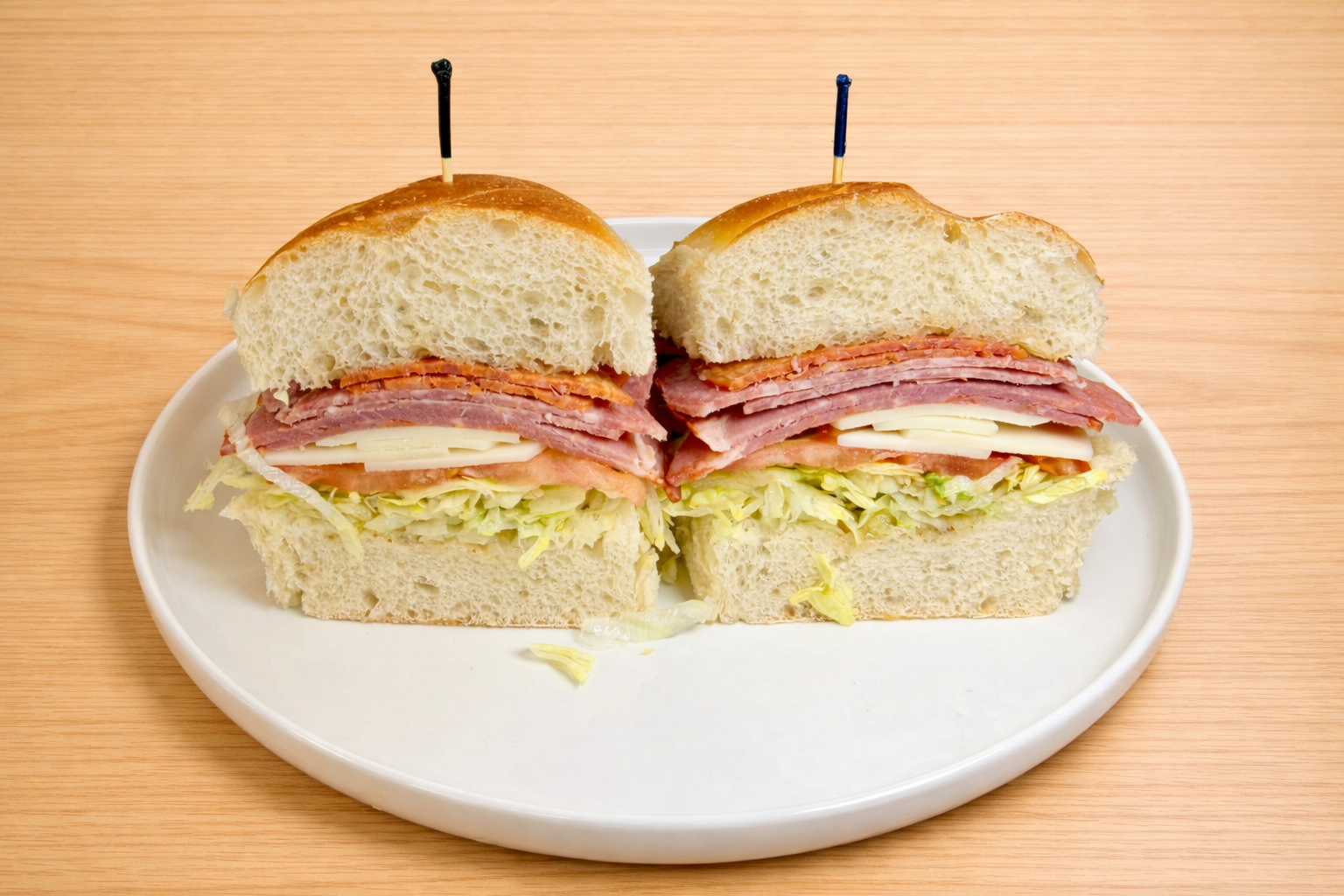 Half-cut sandwich on a white plate, containing lettuce, tomato, cheese, ham, turkey, roast beef, and bacon, with a toothpick on top.