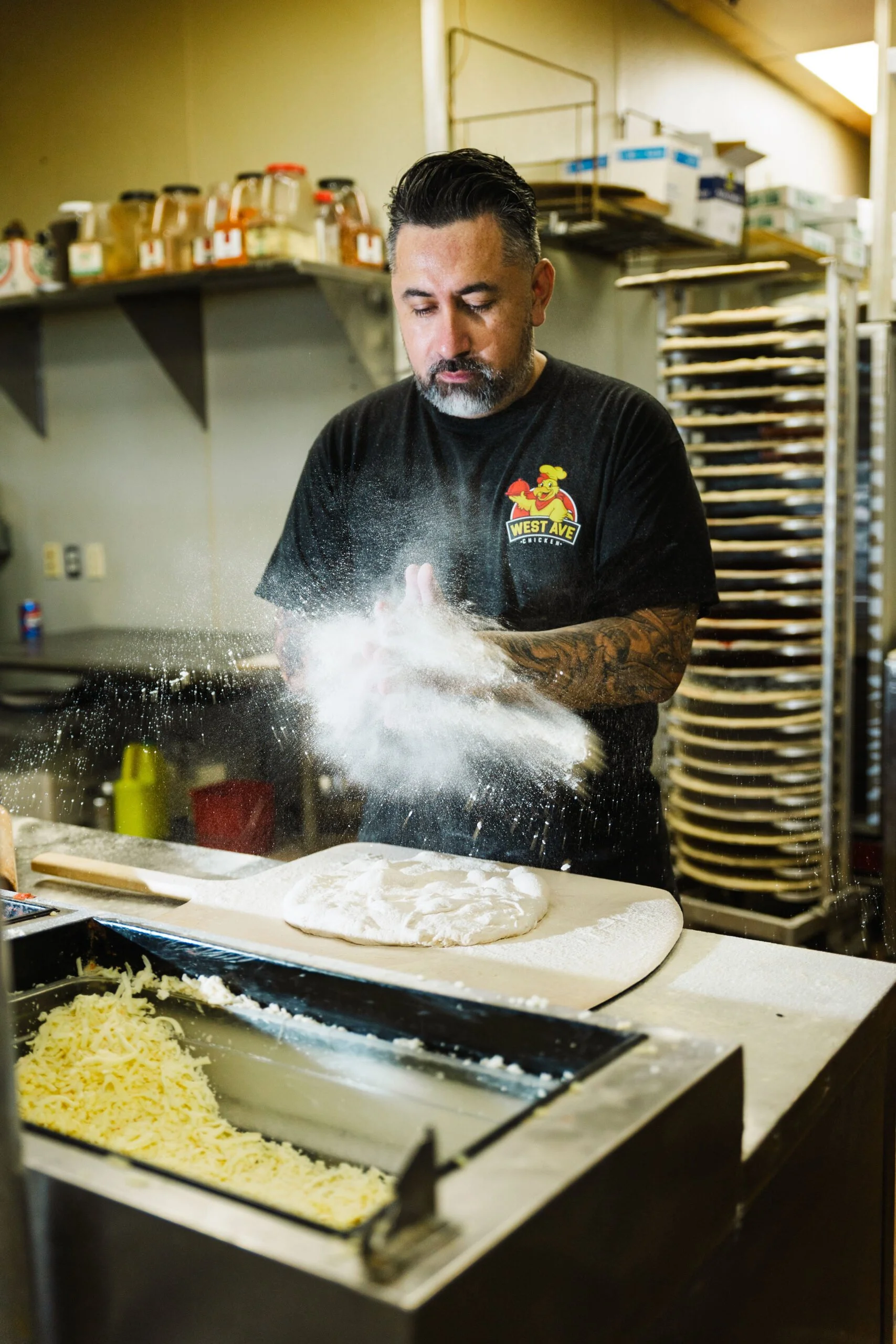A man with black hair and a tattoo on his forearm is tossing flour into the air in a kitchen. He is wearing a black t-shirt with a logo that says 'West Ave.' There is dough on the counter in front of him and shredded cheese in a container nearby. Shelves with jars and trays are visible in the background.