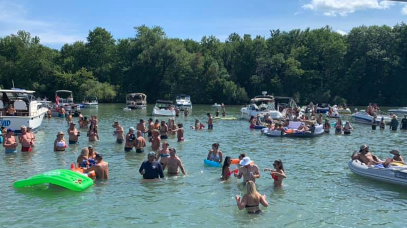 People enjoying a day at the lake, swimming, floating on inflatable tubes, and relaxing on boats under a blue sky with green trees in the background.