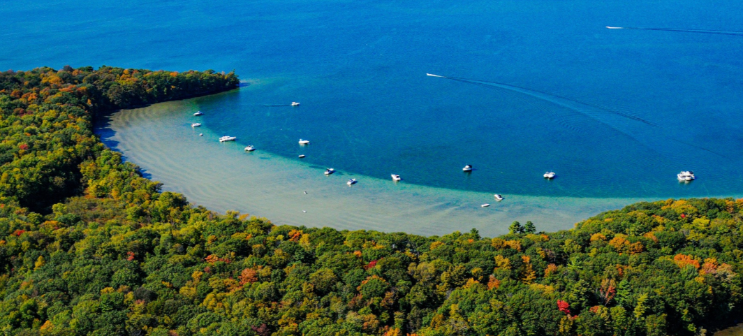 A bay with a curved shoreline surrounded by a dense forest with trees showing fall colors, and several boats anchored in the water with others speeding across the open water.