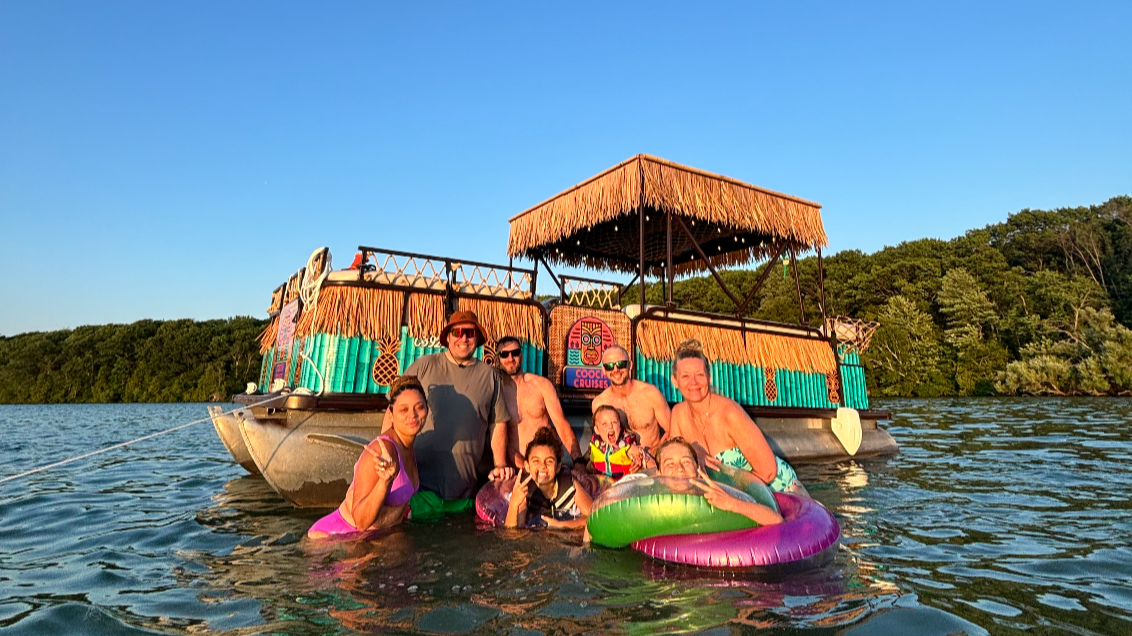 People enjoying a boat party with a floating dock and a tiki hut in a lake during sunset.