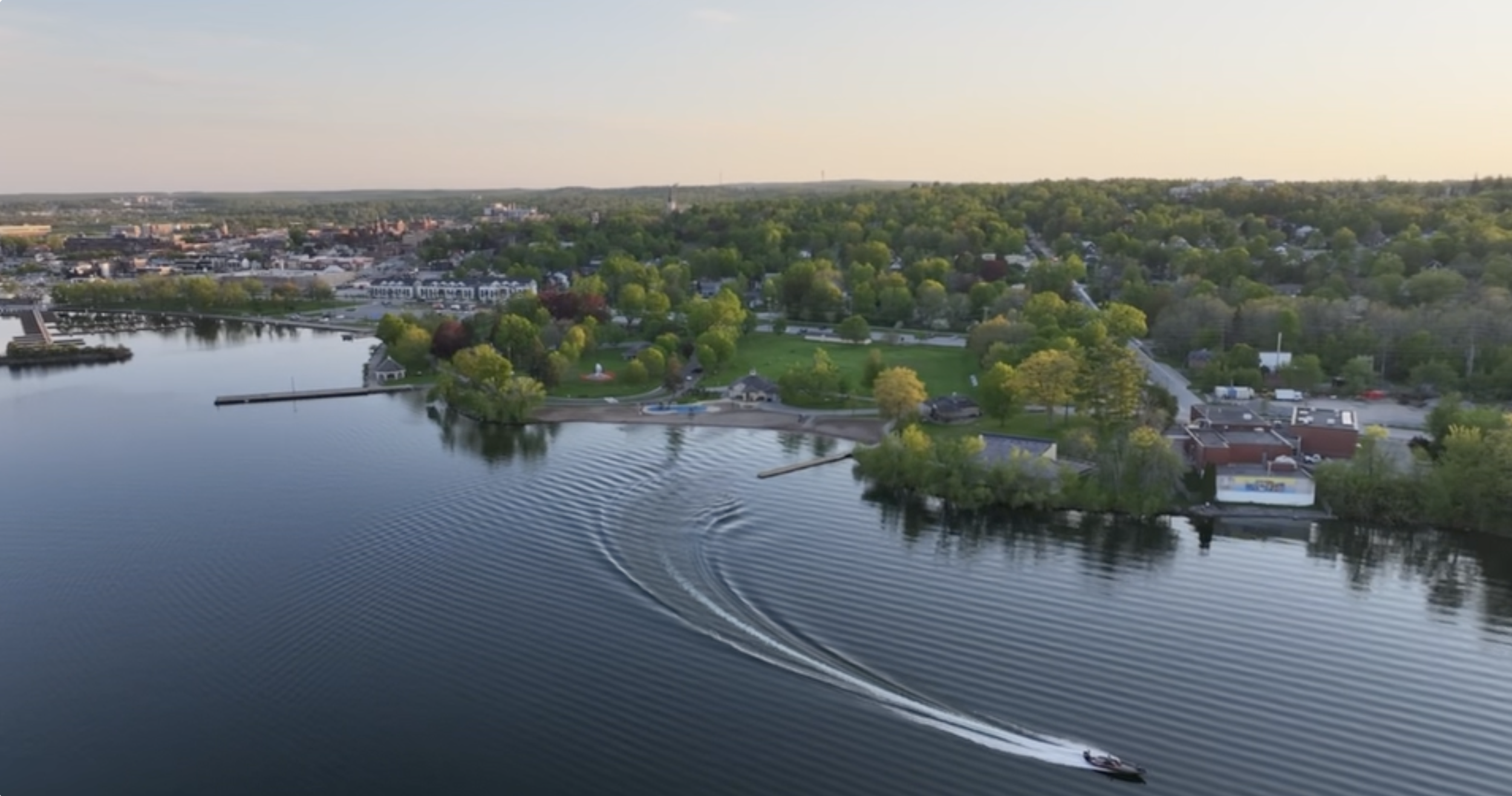 Aerial view of a town with a lake, green park, and cityscape in the background during sunset.