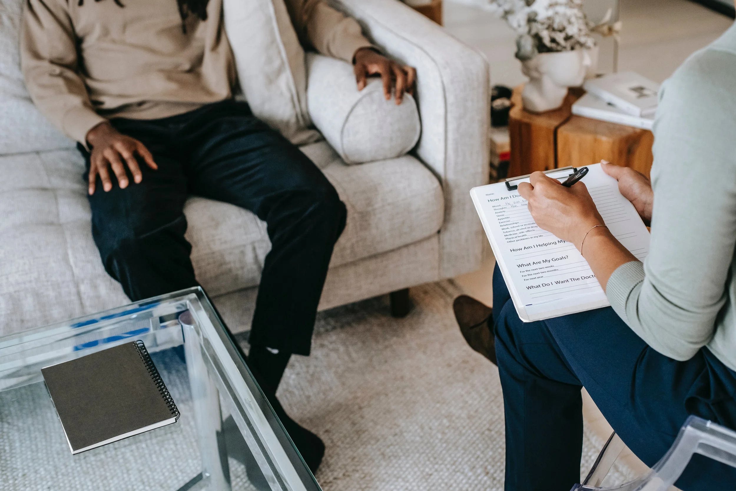 A person sitting on a beige couch during a therapy session, with a therapist holding a clipboard. The scene takes place in a living room with a small table, books, a decorative bust, and a vase with flowers in the background.