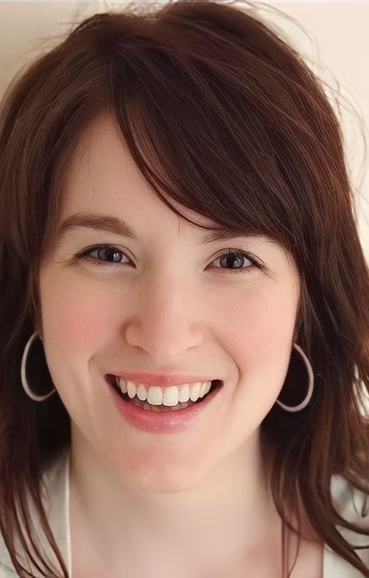 Close-up of a woman smiling with brown hair, wearing hoop earrings, showing teeth.