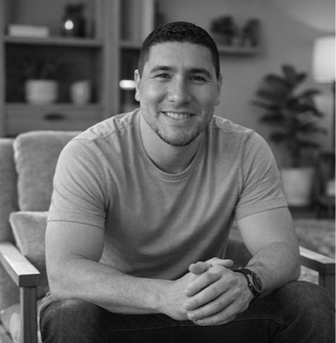 A smiling man sitting on a couch in a living room with shelves and plants in the background, black and white photo.