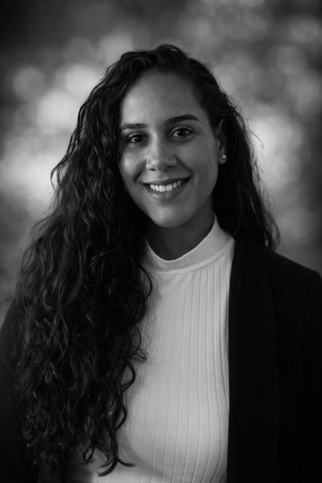 A young woman with long, curly hair smiling at the camera, wearing a white high-neck top and a dark blazer, in black and white.