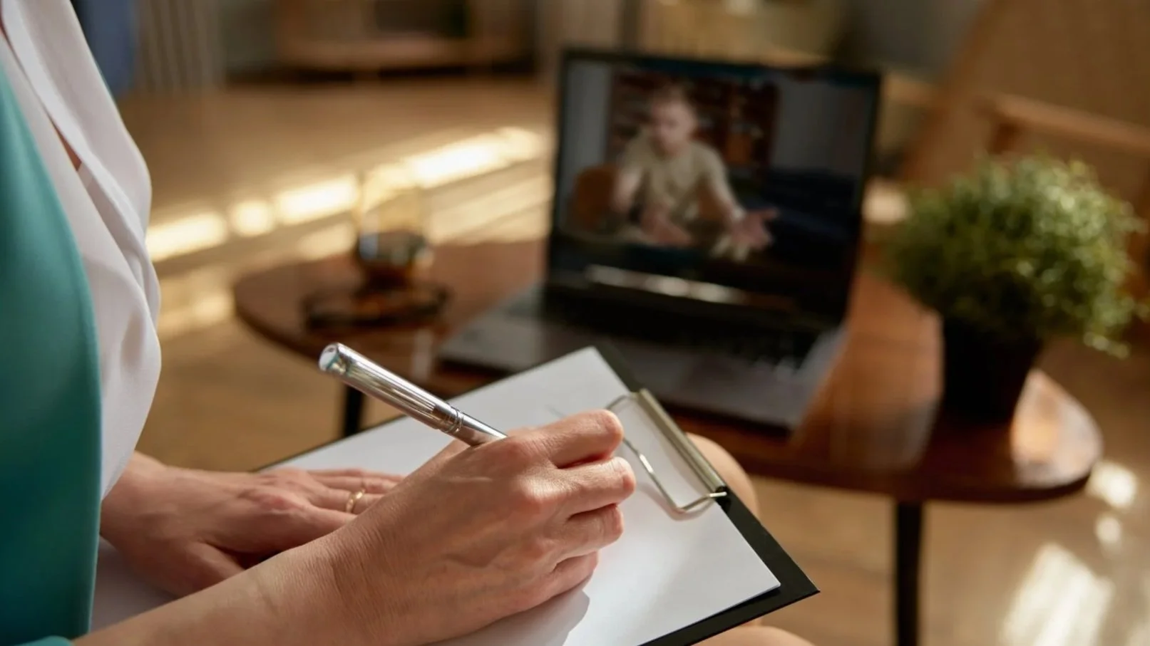 Close-up of a person writing in a notebook with a roller pen, sitting in front of a laptop displaying a video call with a young woman. A plant and a glass of beverage are on a table in the background.
