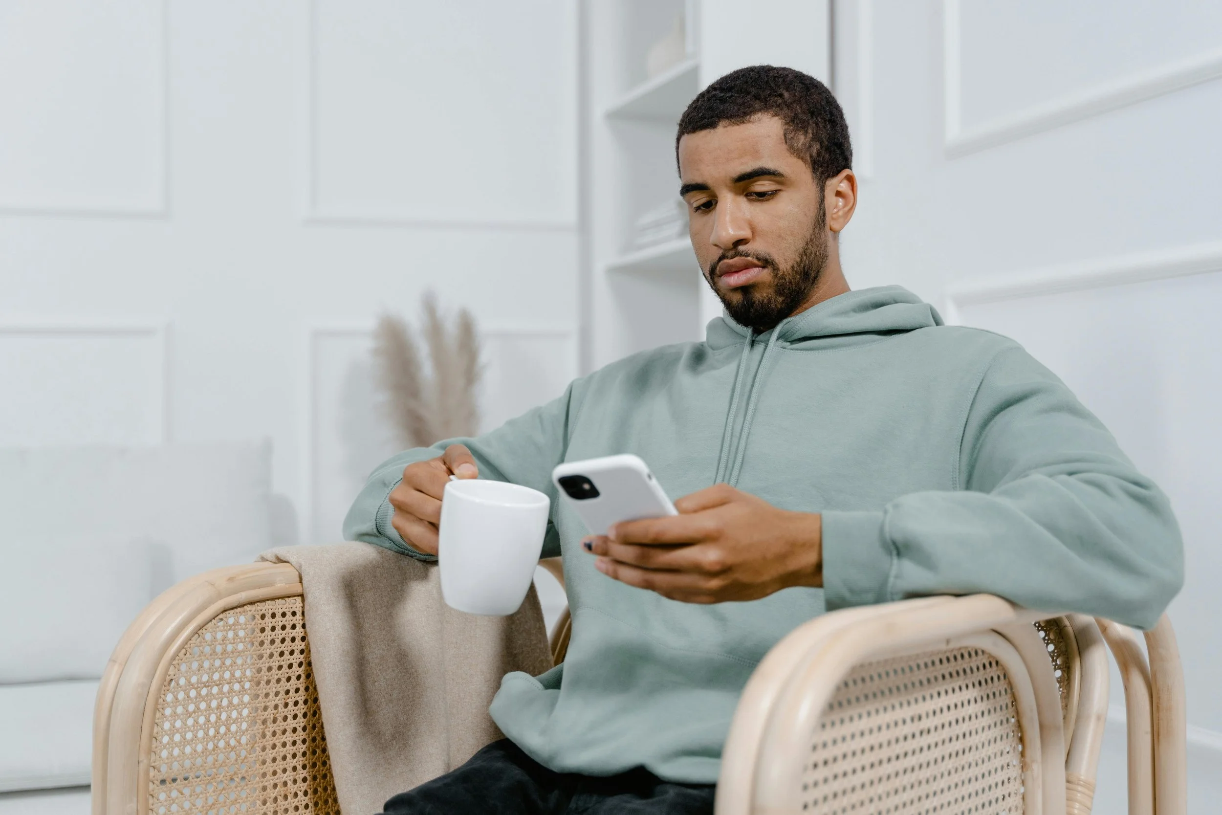 A young man with short dark hair and a beard, wearing a light gray hoodie, sits in a wicker chair holding a smartphone in his right hand. There is a white mug and a beige blanket on the arm of the chair. The background features white walls with simple panel detailing and a shelf with minimal decor.