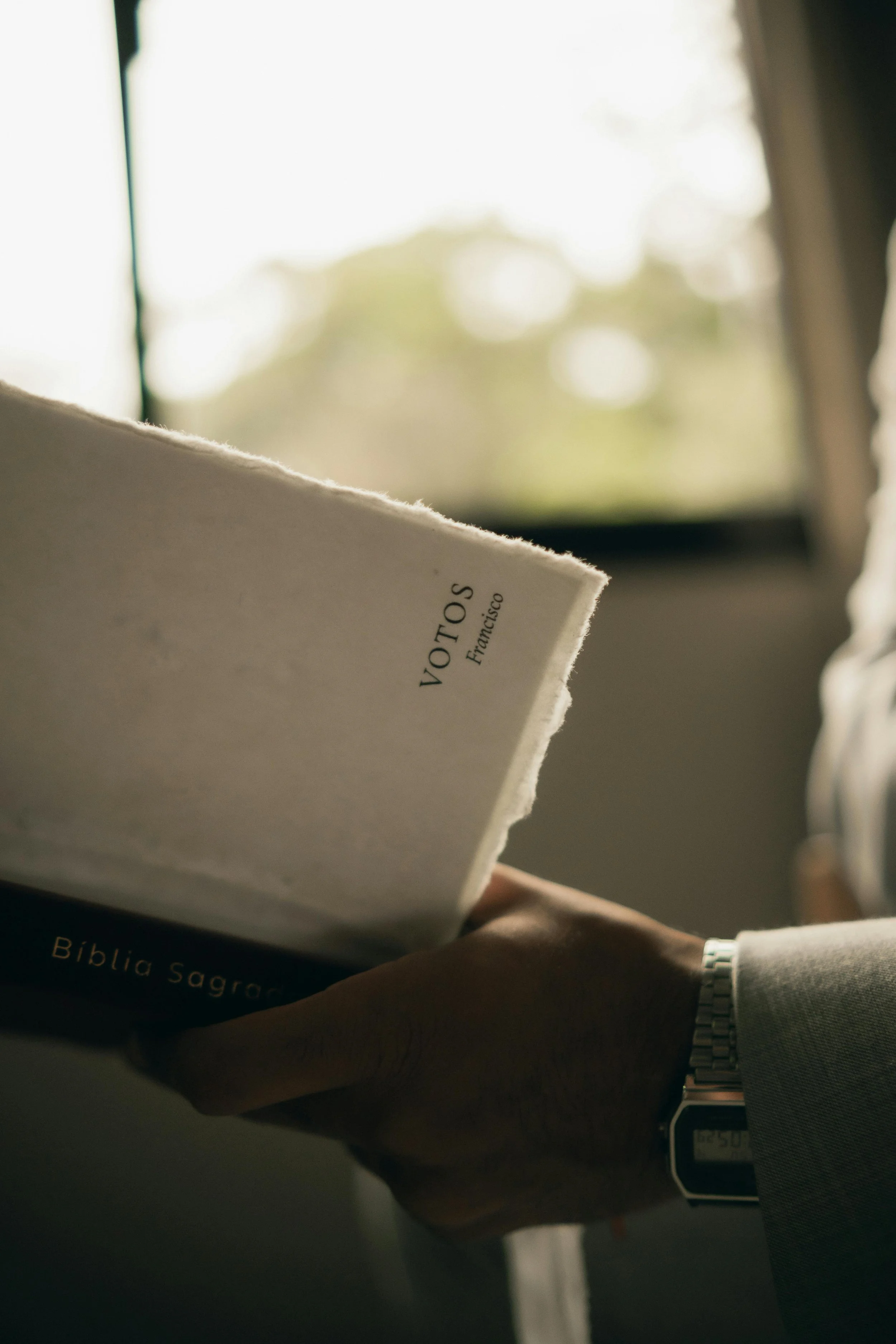 A person wearing a watch on their left wrist is holding a Bible with the title 'Bíblia Sagrada' in Portuguese, near a window with sunlight coming through.