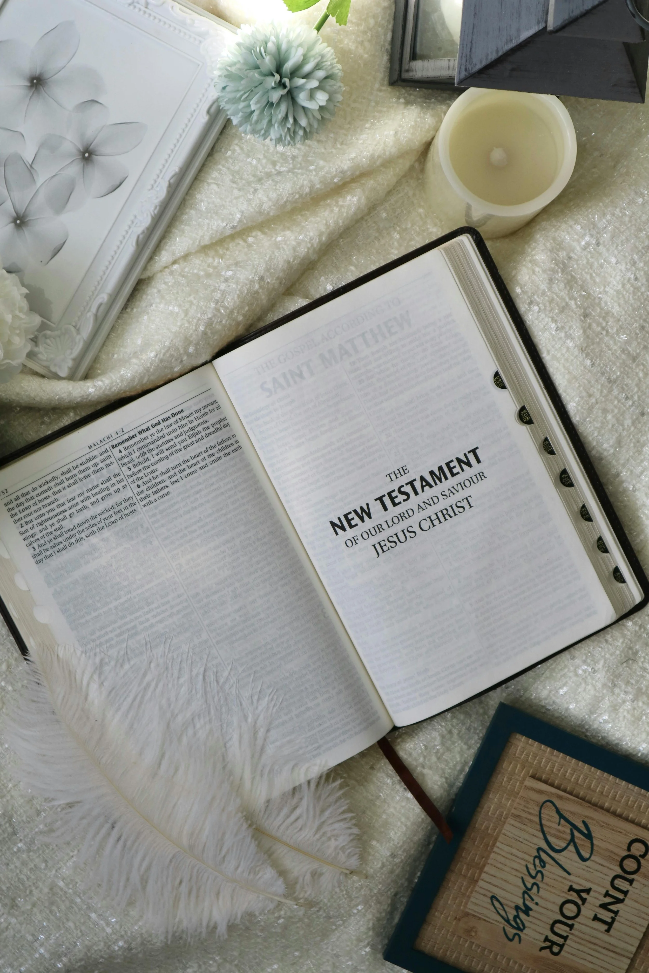 Open Bible with the title 'The New Testament' on a lace tablecloth, beside a white feathered decoration, a white candle, a flower arrangement, and framed decorative items.