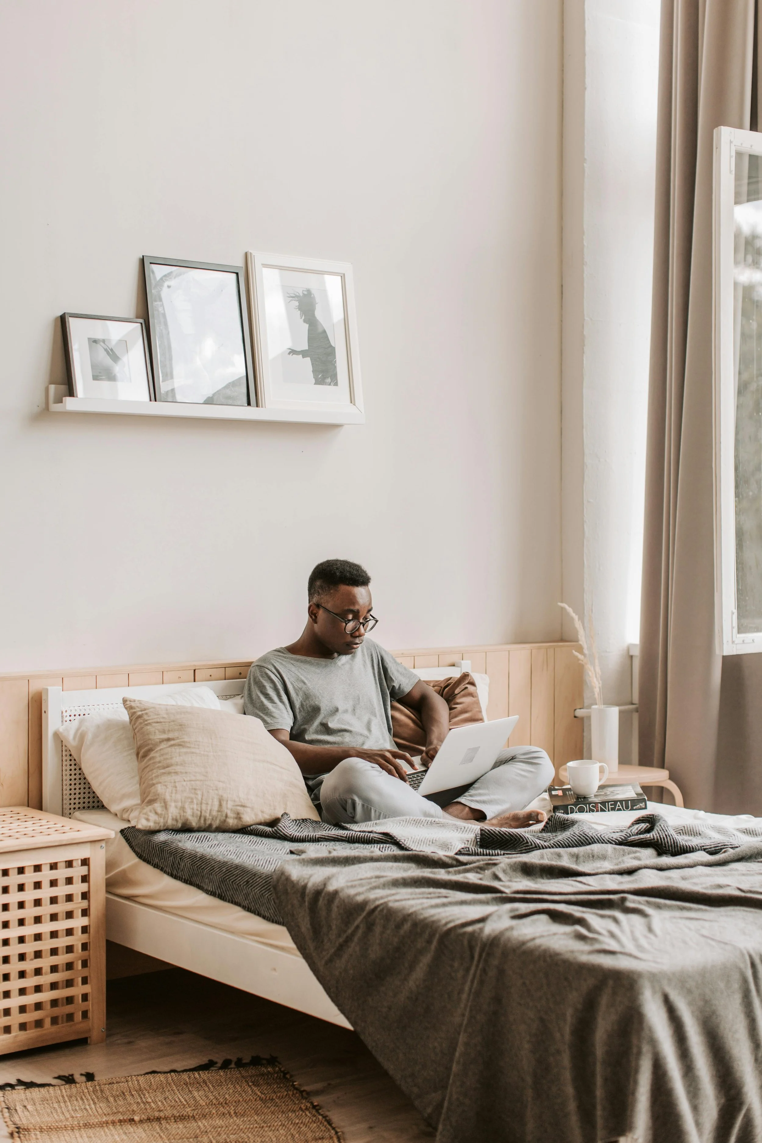 A man sits on a bed using a laptop, with a window and beige curtains behind him. The bed has beige pillows and a gray blanket. A small nightstand with a white mug, a book titled 'POIS NEAU,' and some papers is beside the bed. Three framed black and white silhouettes hang above the bed.