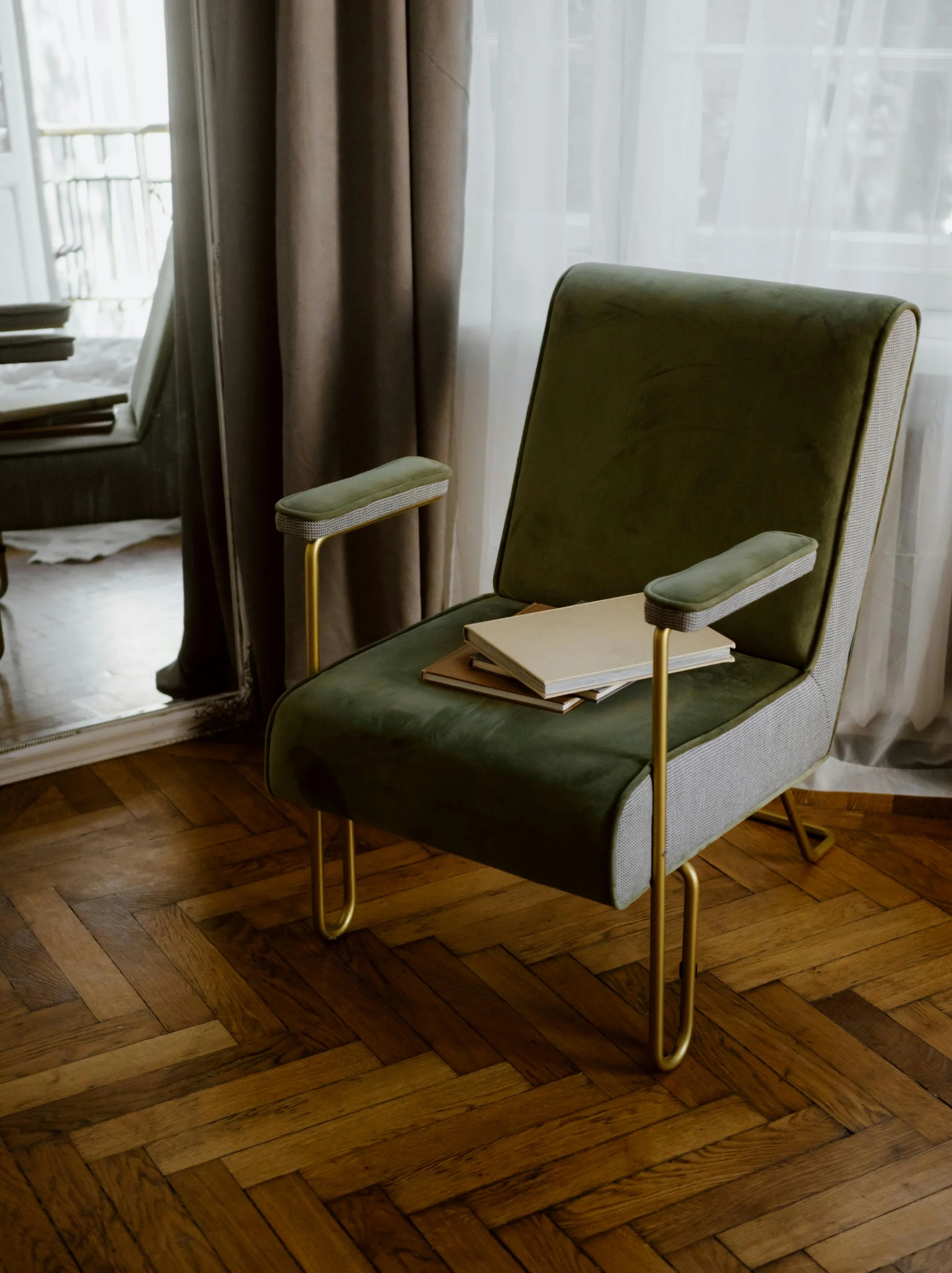 A green armchair with gold metal legs, placed next to a table with notebooks on it, in front of a window with sheer curtains.