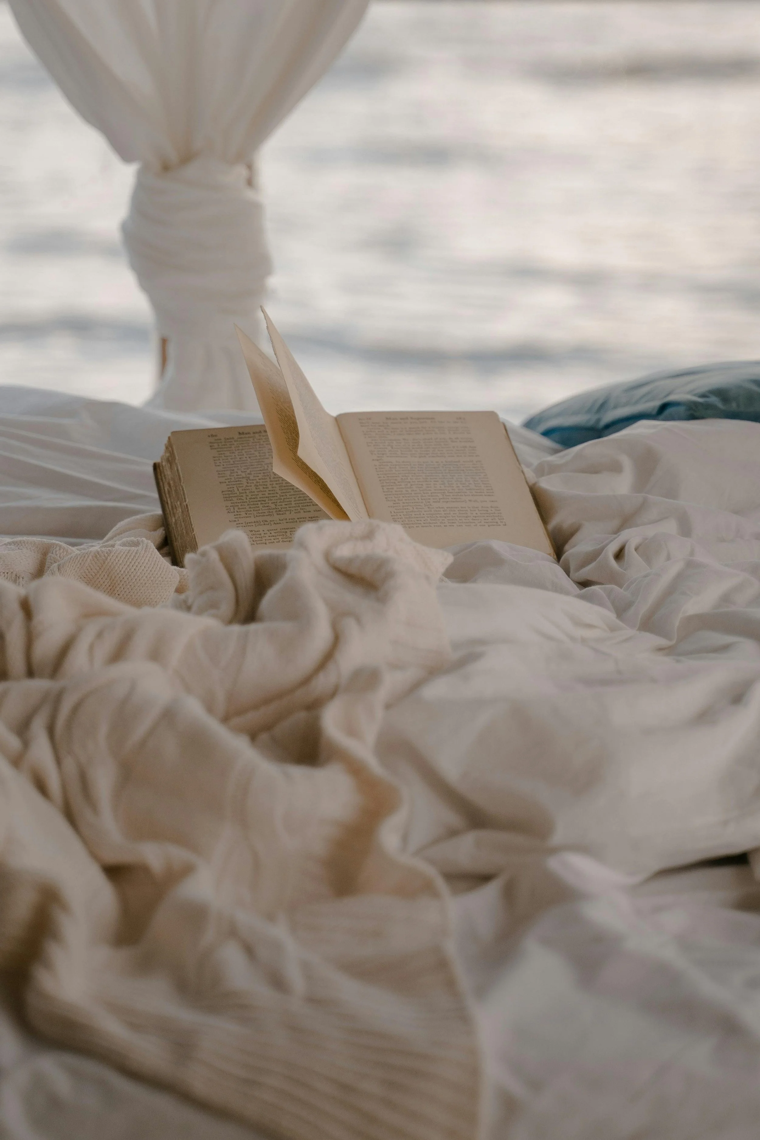 A book resting on cream-colored bed linens with a background of water, possibly an ocean or lake, seen through a window or opening.