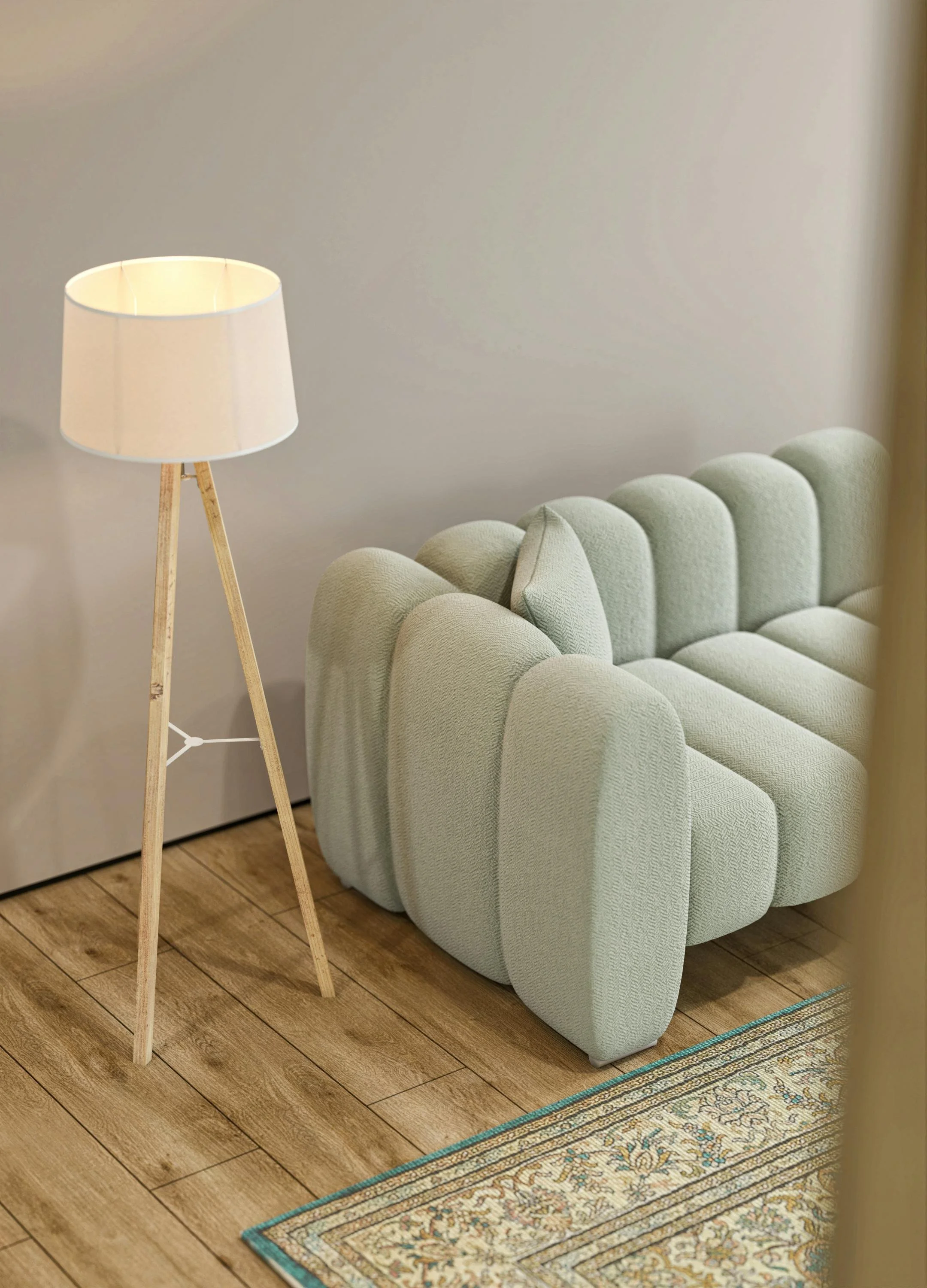 Close-up of a light green upholstered sofa with rounded armrests next to a wooden floor, a patterned rug, and a tall wooden tripod floor lamp with a white lampshade.