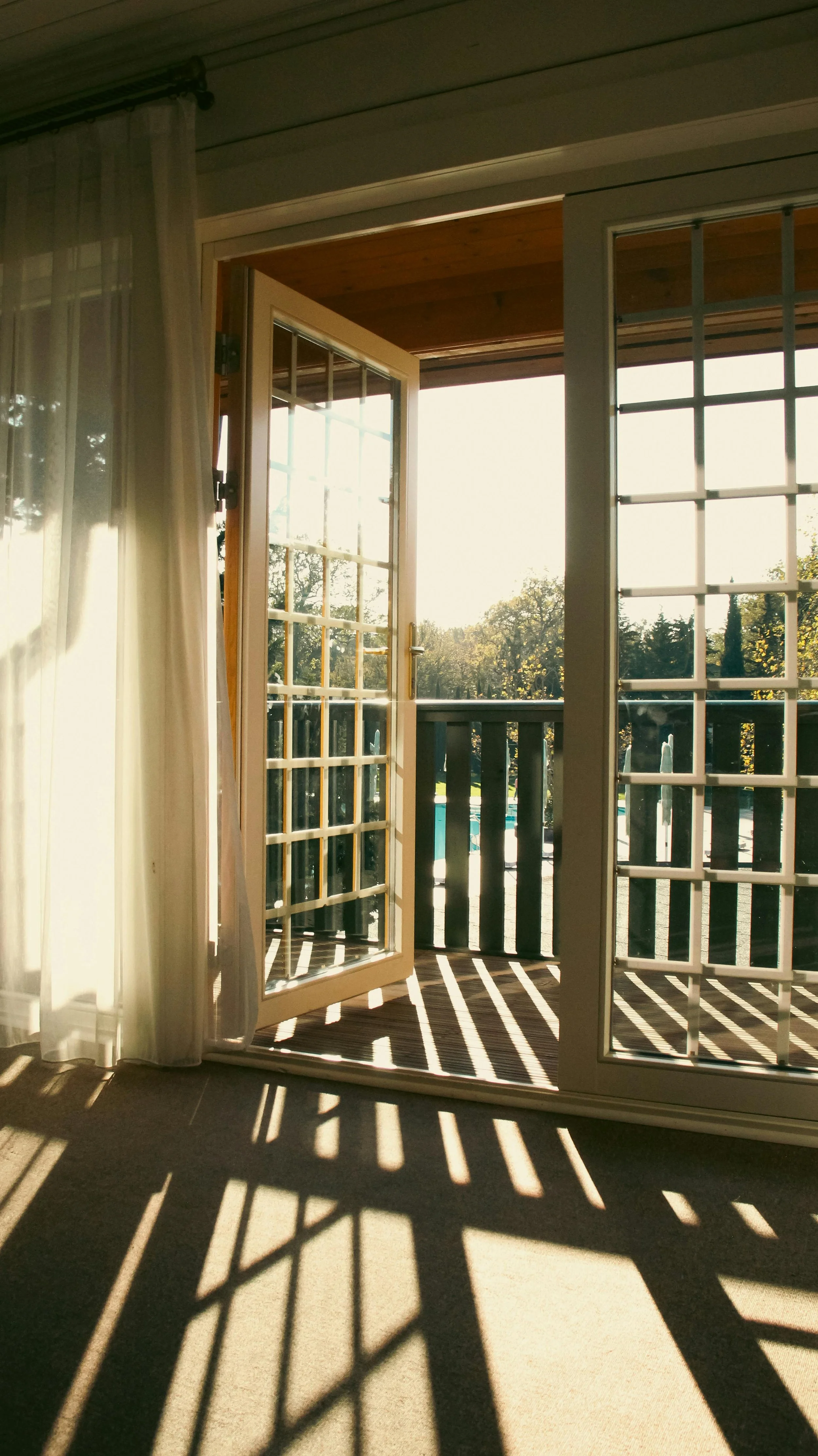 Open French doors leading to a balcony with a railing, overlooking trees with golden leaves in sunlight, casting shadows inside a room.