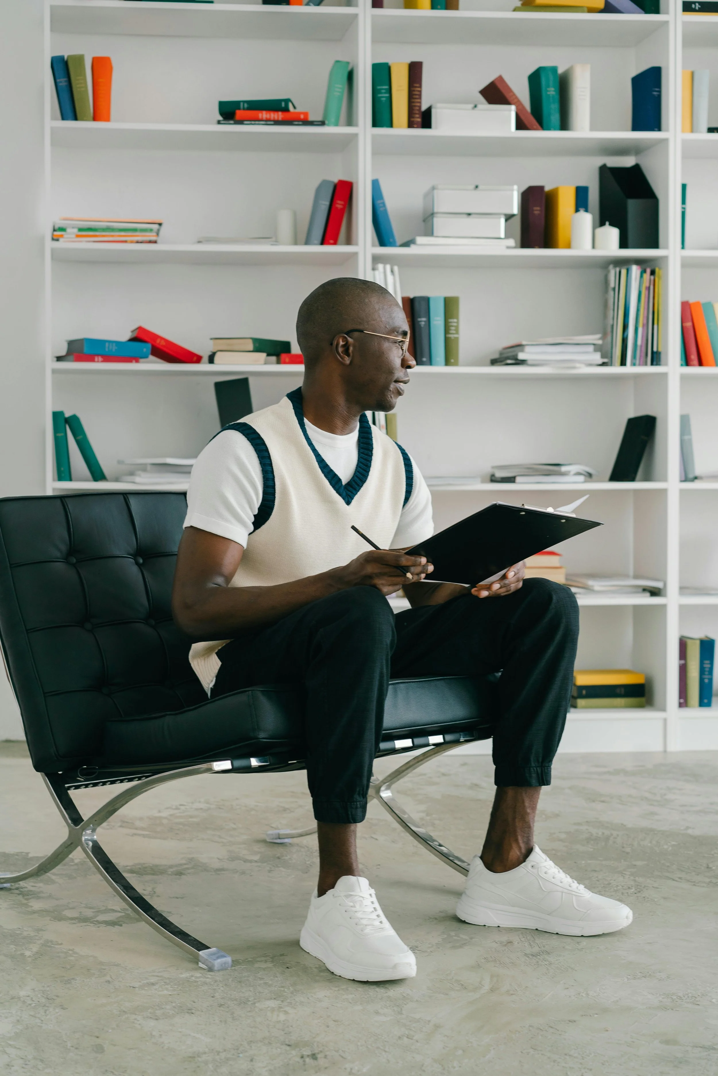 A man sitting on a black leather lounge chair with metal frame, holding a clipboard and pen, wearing a white vest with dark trim over a white t-shirt, black pants, and white sneakers, in front of a white bookshelf filled with colorful books and binders.
