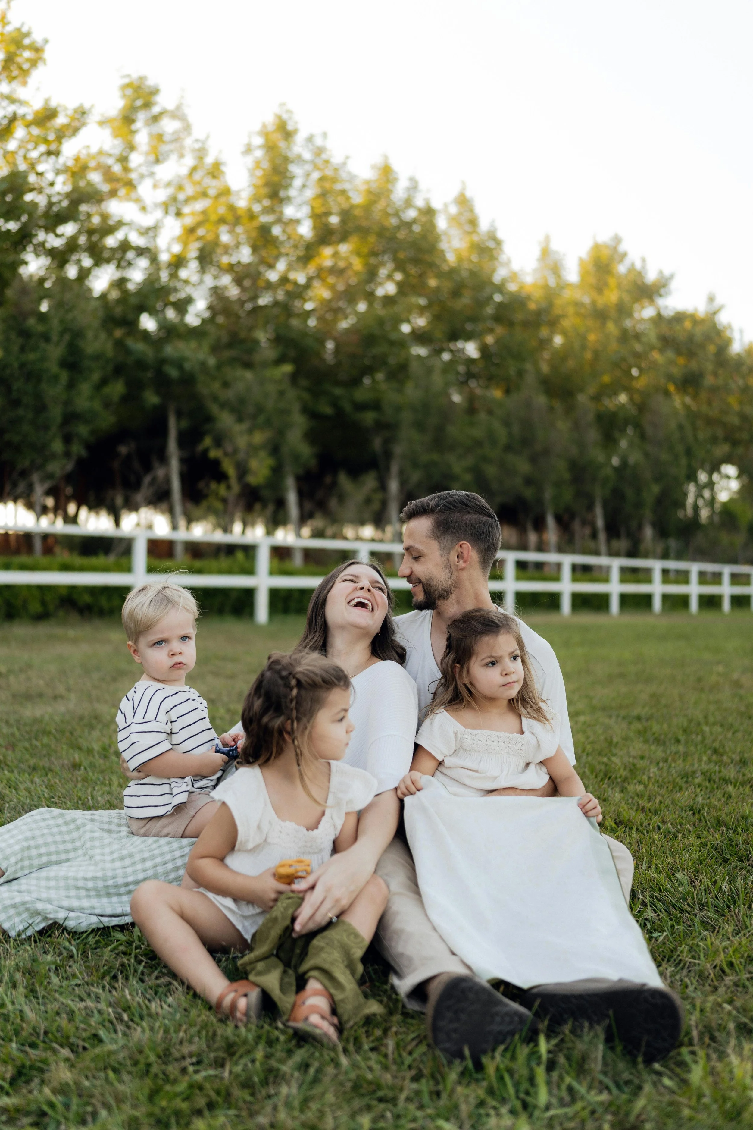 A family of six sitting on a blanket in a grassy area outdoors, smiling and interacting, with a background of trees and a white fence.
