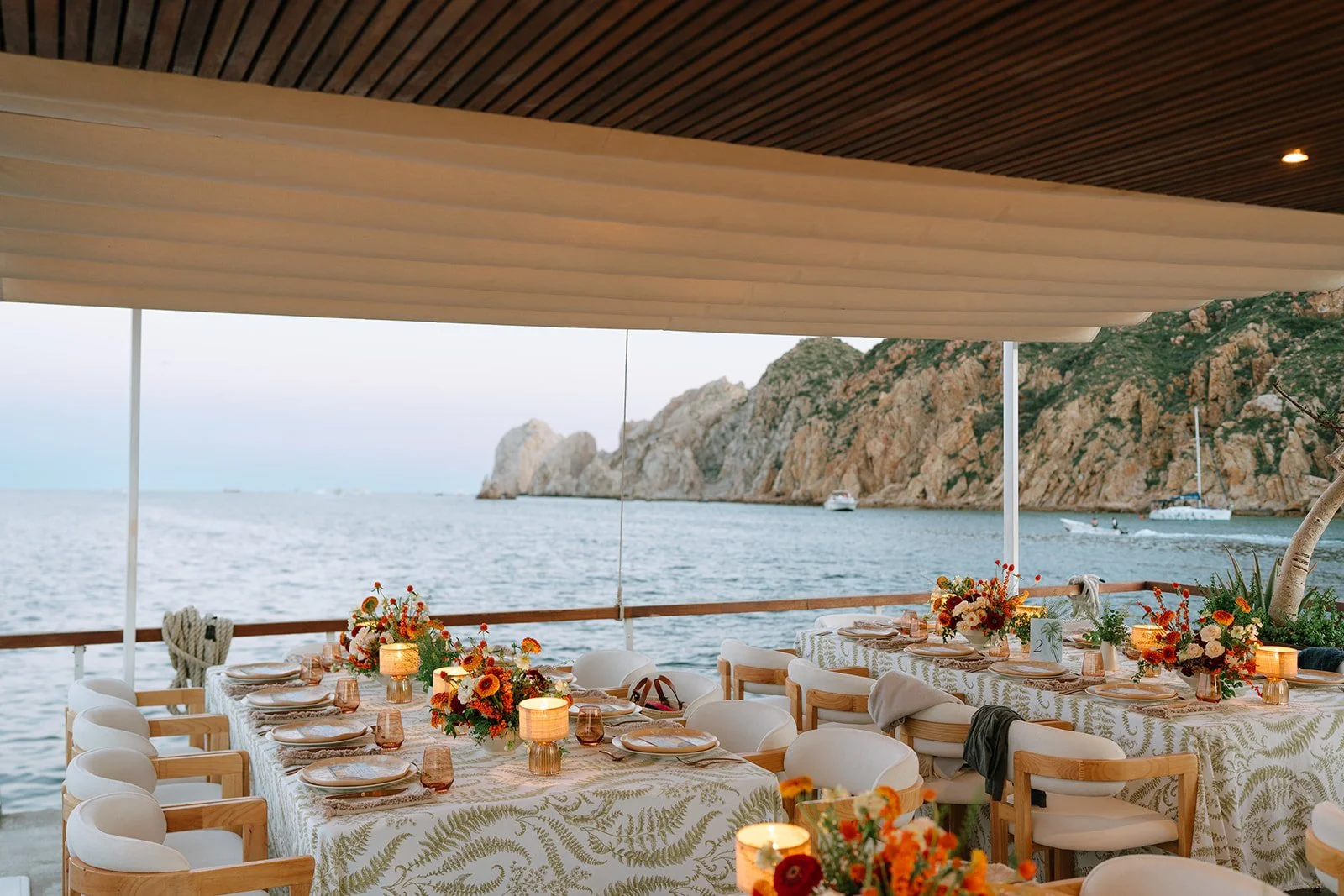 Set outdoor dining table overlooking a body of water with boats and rocky cliffs in the background. The table is decorated with floral arrangements, tableware, and candles.