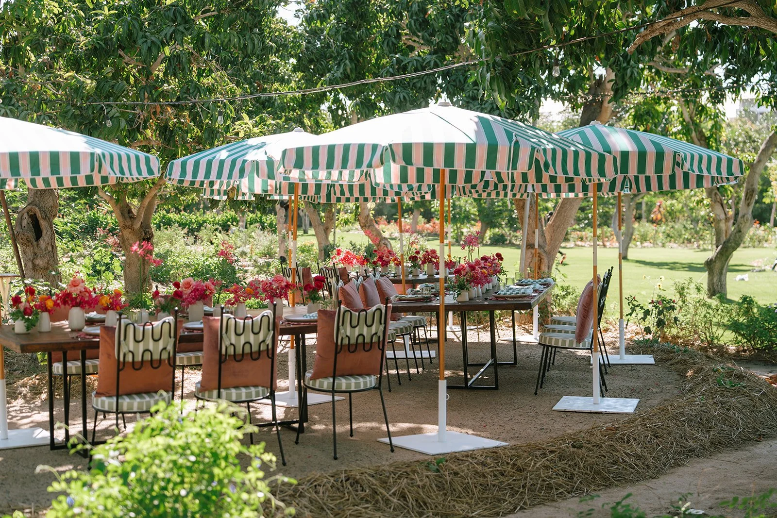 Outdoor dining setup with tables, chairs, and pink flower arrangements under large green and white striped umbrellas in a garden.