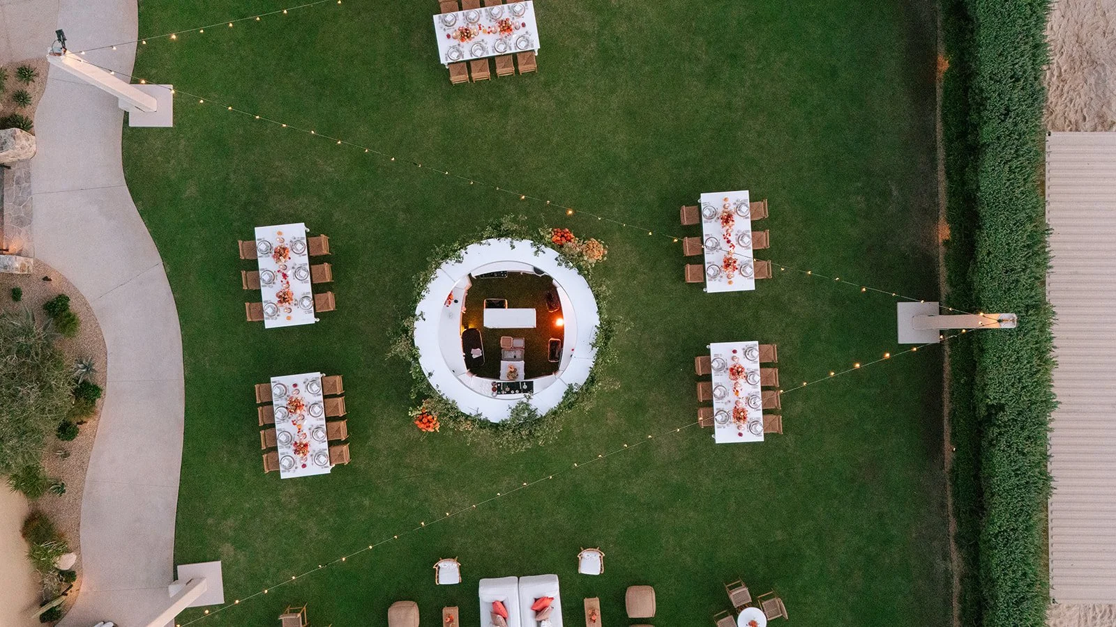Aerial view of an outdoor dinner party setup on a grassy yard with six rectangular tables decorated with orange and red floral centerpieces, string lights hanging across the yard, and a circular white lounge area with chairs and a table in the center.